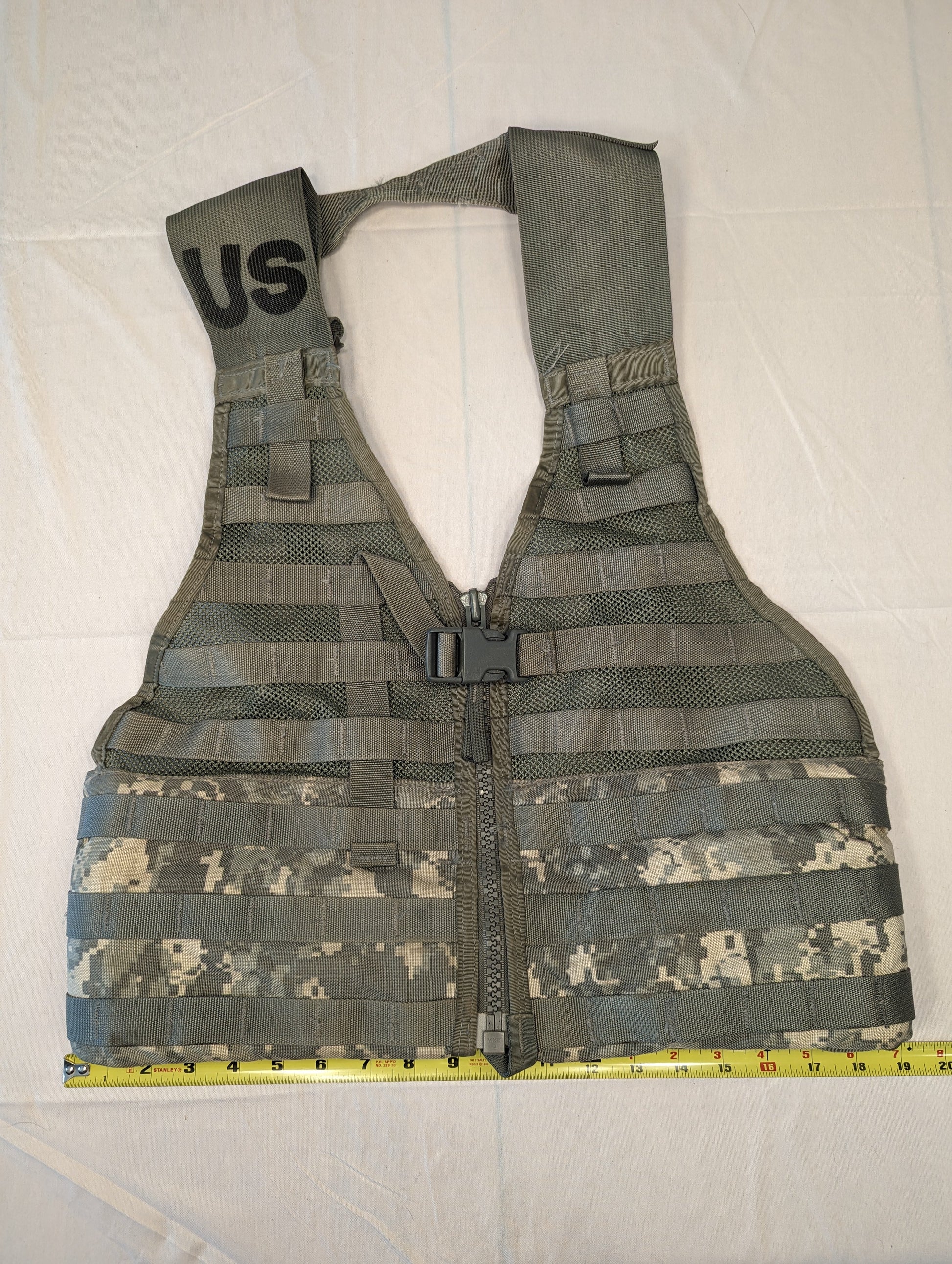 A U.S. Military Surplus Load Carrier Vest (FLC) in ACU digital camouflage and olive green, laid flat with a tape measure showing its width, and featuring the stenciled 'US' mark on the shoulder strap.