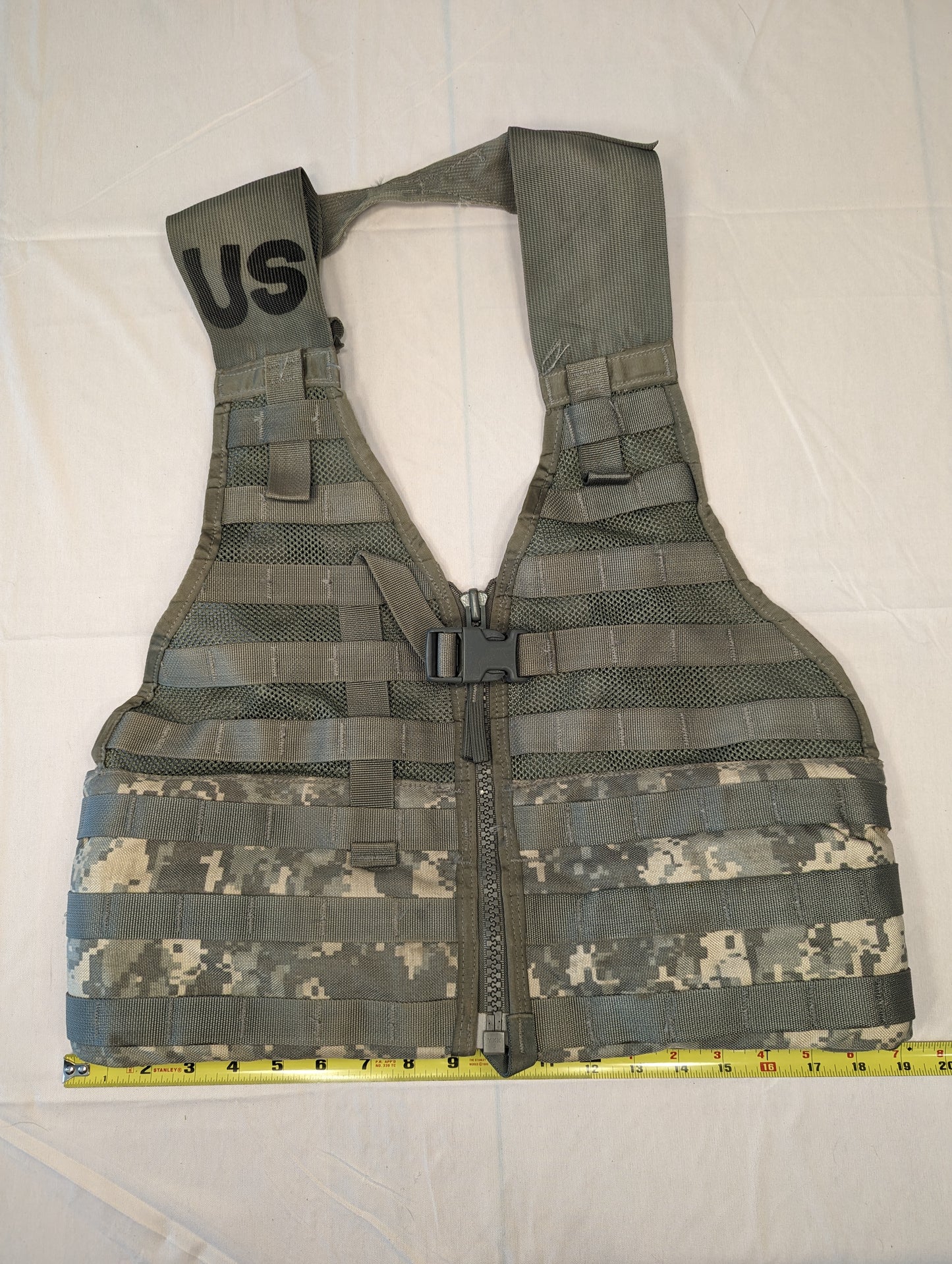 A full front view of a U.S. Military Surplus Load Carrier Vest (FLC) in ACU digital camouflage, unaccessorized, laid flat with a tape measure showing its width, and featuring the large stenciled 'US' mark.
