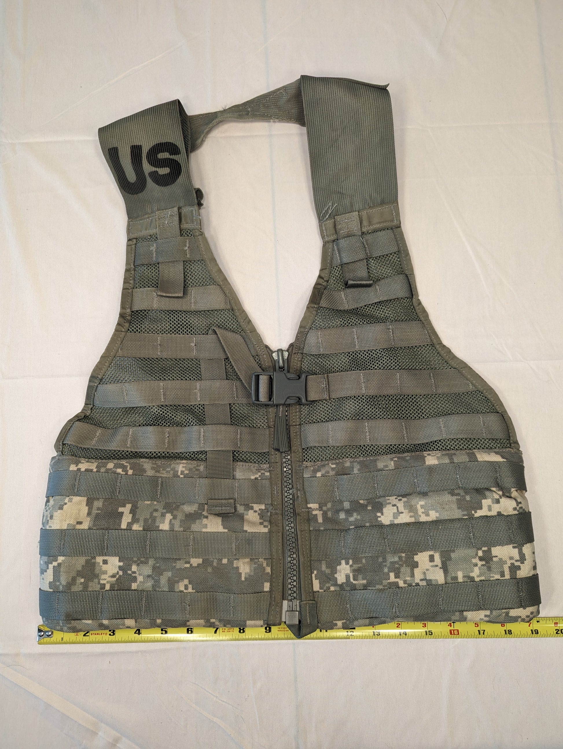 A full front view of a U.S. Military Surplus Load Carrier Vest (FLC) in ACU digital camouflage, unaccessorized, laid flat with a tape measure showing its width, and featuring the large stenciled 'US' mark.