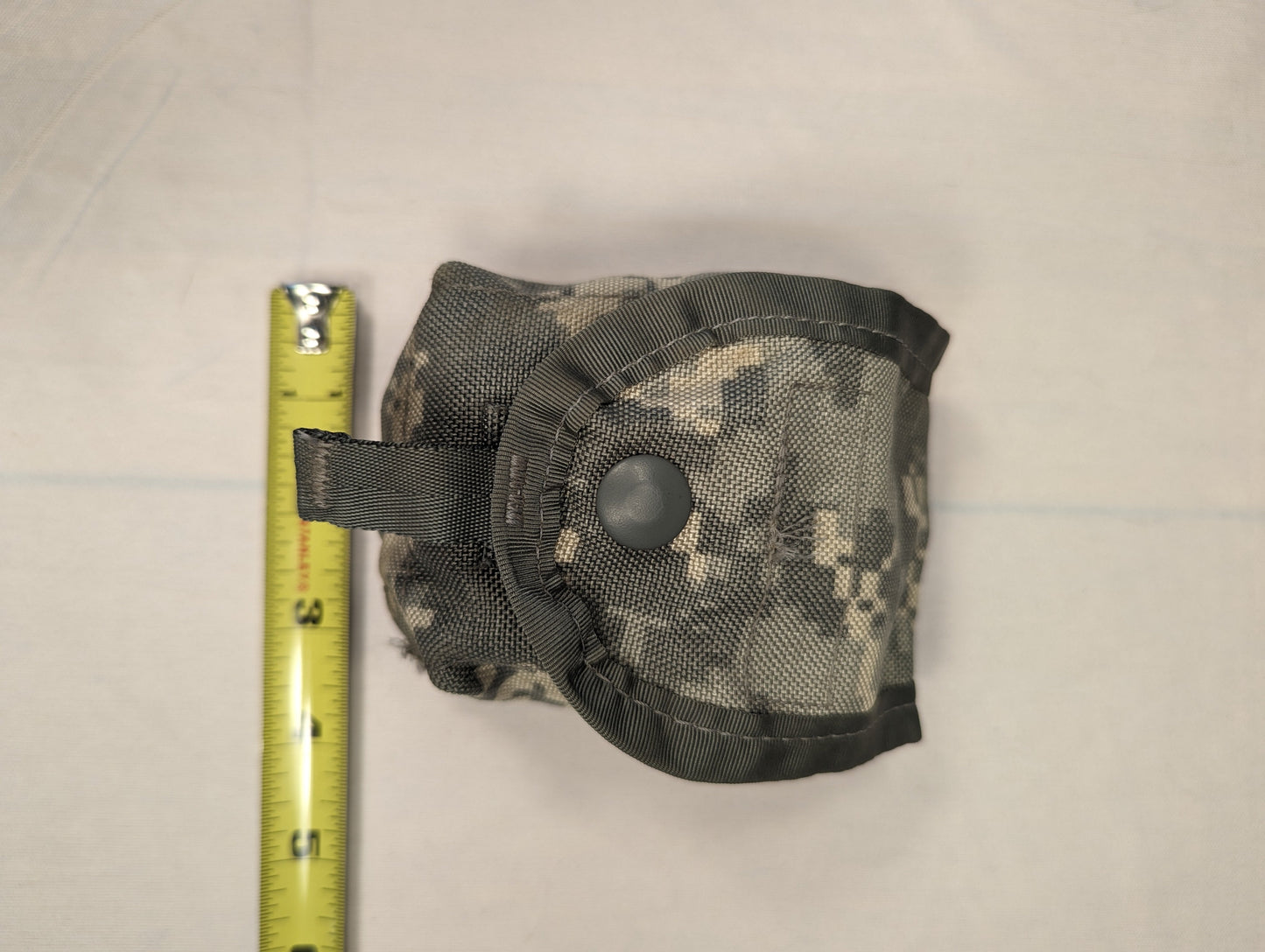 A tightly rolled U.S. Military Surplus accessory pouch in ACU digital camouflage, secured with a snap button closure and photographed with a measuring tape to indicate its compact size.
