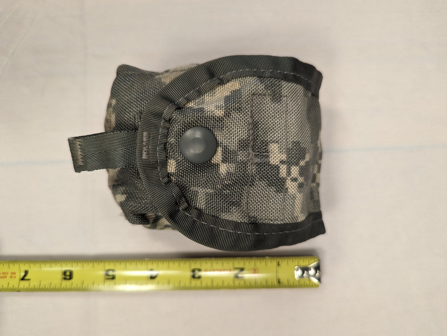 A tightly rolled U.S. Military Surplus accessory pouch in ACU digital camouflage, secured with a snap button closure and photographed with a measuring tape to indicate its compact size.