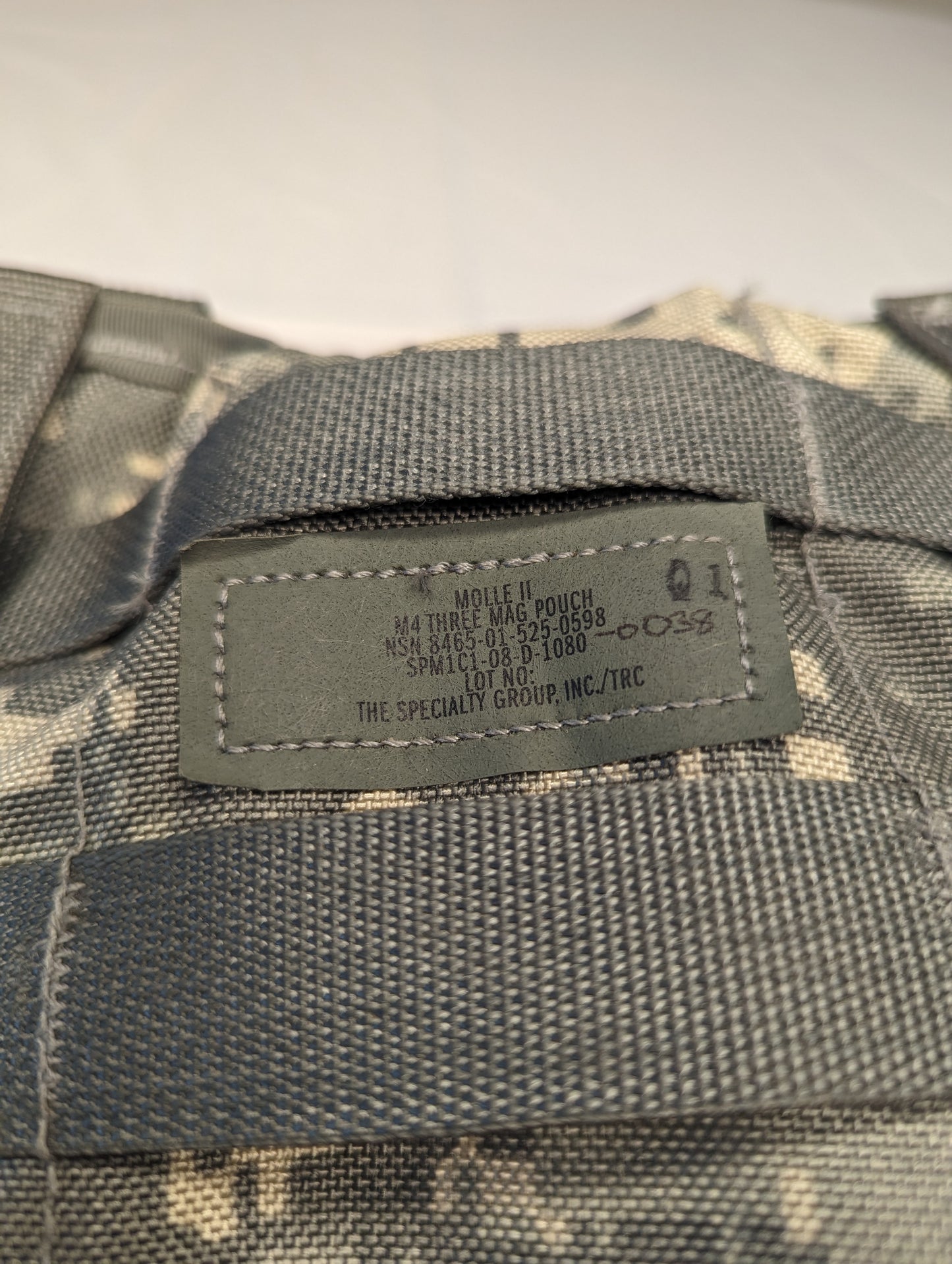 A close-up photograph of the official U.S. Military issue tag sewn onto the ACU digital camouflage MOLLE webbing, displaying the product name 'POUCH, TRIPLE MAGAZINE', NSN 8465-01-525-0589, and manufacturer 'THE SPECIALTY GROUP, INC.'.