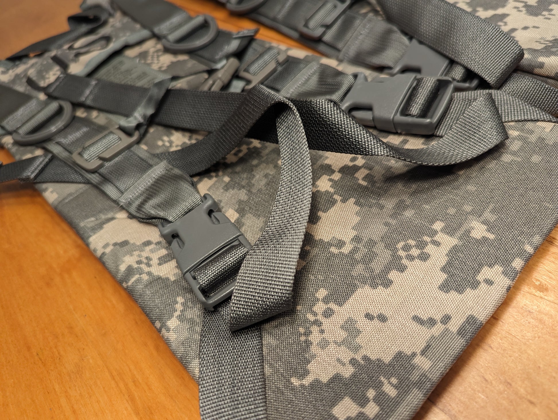 A close-up detail shot of the adjustment system on a U.S. Military ACU pack, featuring numerous black plastic quick-release buckles and webbing straps over the digital camouflage fabric.