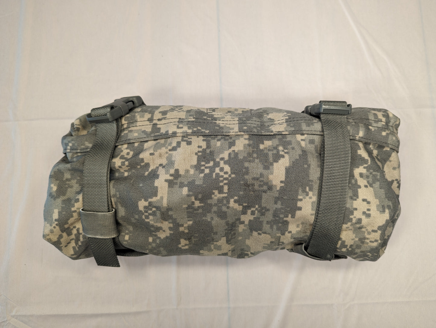 A tightly rolled U.S. Military Surplus camouflage gear pouch or carrier in ACU digital camouflage, secured by two black webbing straps with quick-release buckles, resting horizontally.