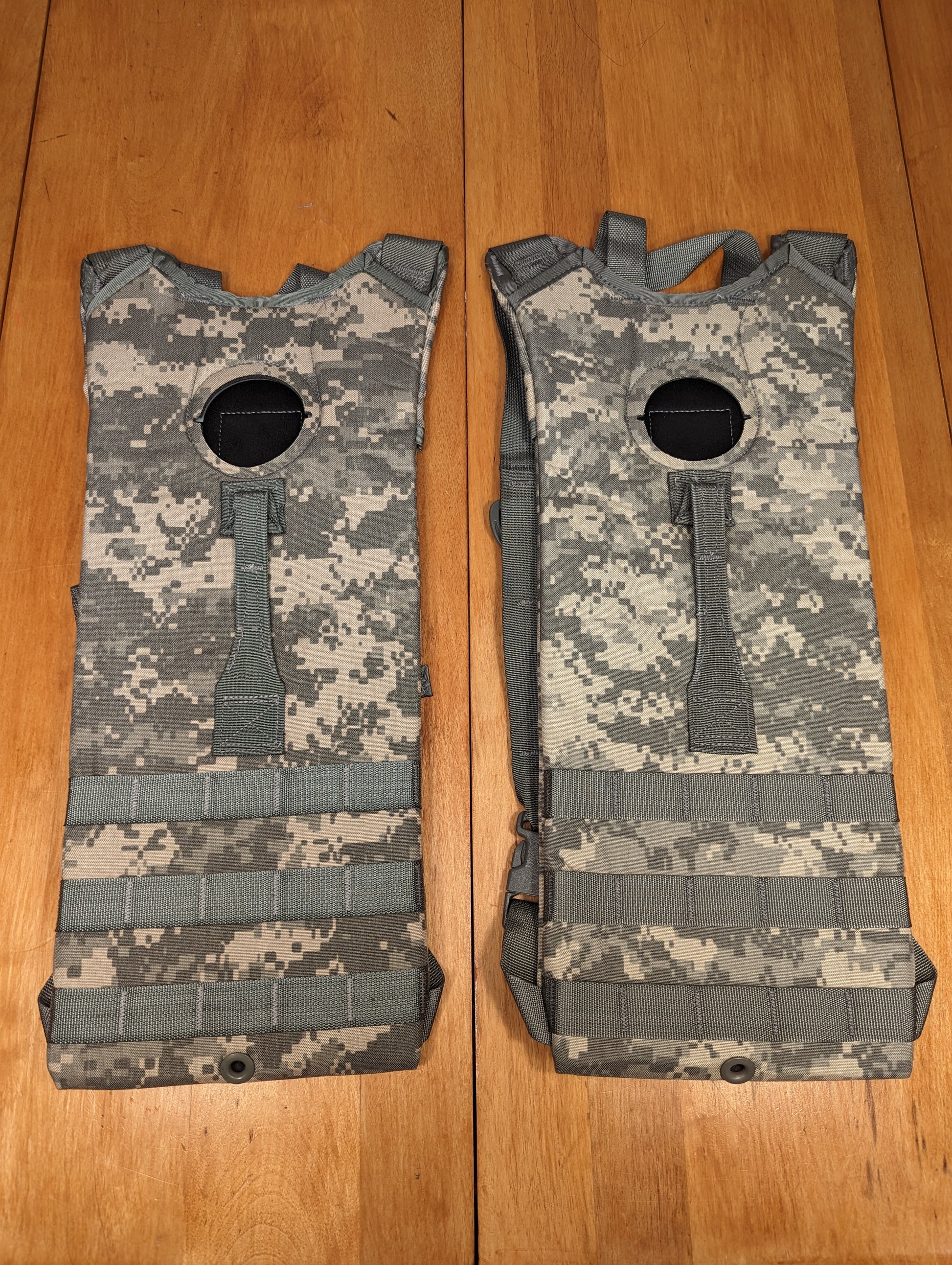 A pair of U.S. Military Surplus Hydration Carriers in ACU/UCP digital camouflage, laid side-by-side on a wooden surface, showing the front panel with horizontal MOLLE webbing and the top filler port opening.