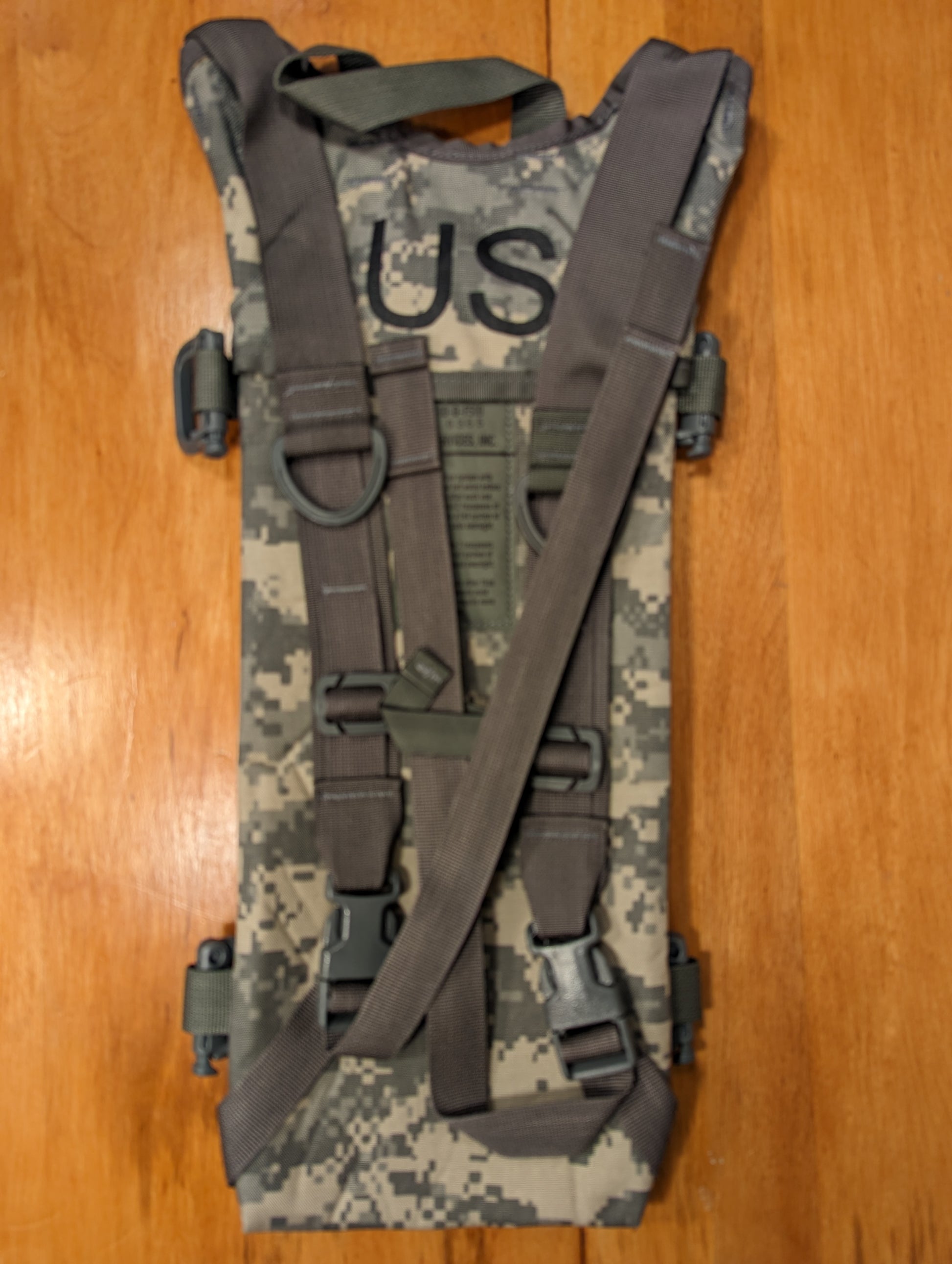 A back view of a U.S. Military Hydration Carrier in ACU digital camouflage, clearly displaying the padded shoulder harness, adjustment webbing, large D-rings, and the stenciled 'US' government mark.