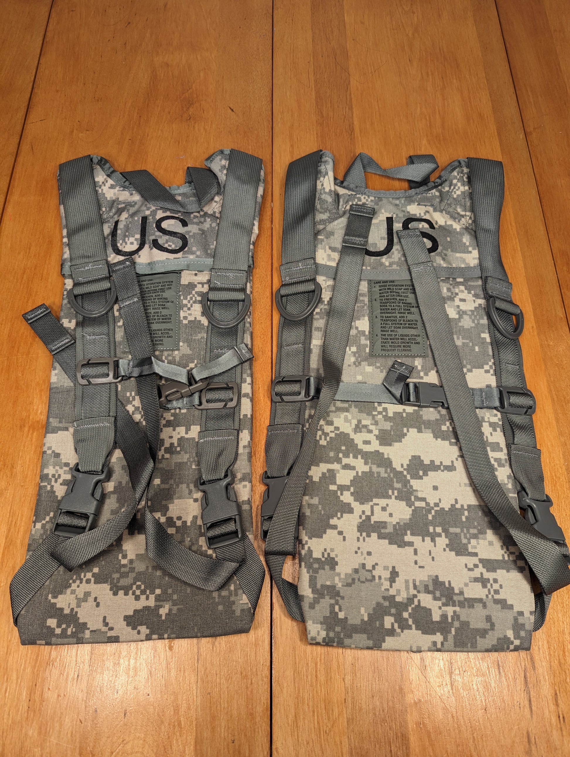 A pair of new U.S. Military Hydration Carrier harnesses in ACU digital camouflage, laid side-by-side on a wooden surface, clearly displaying the padded shoulder straps, adjustment webbing, and the stenciled 'US' mark on each pack.