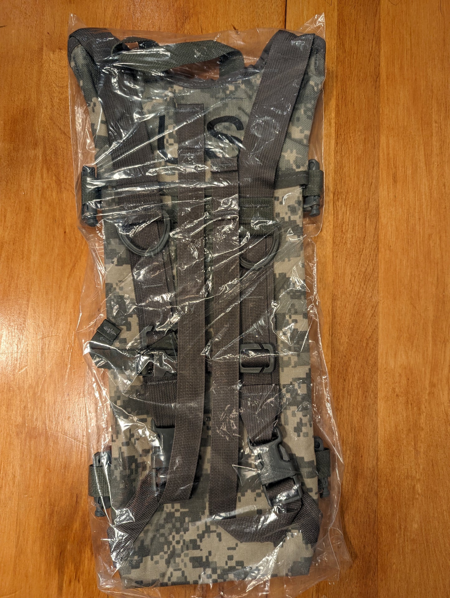 A new U.S. Military Hydration Carrier in ACU/UCP digital camouflage, sealed inside clear plastic packaging, showing the complete shoulder harness, chest strap, and black quick-release buckles.