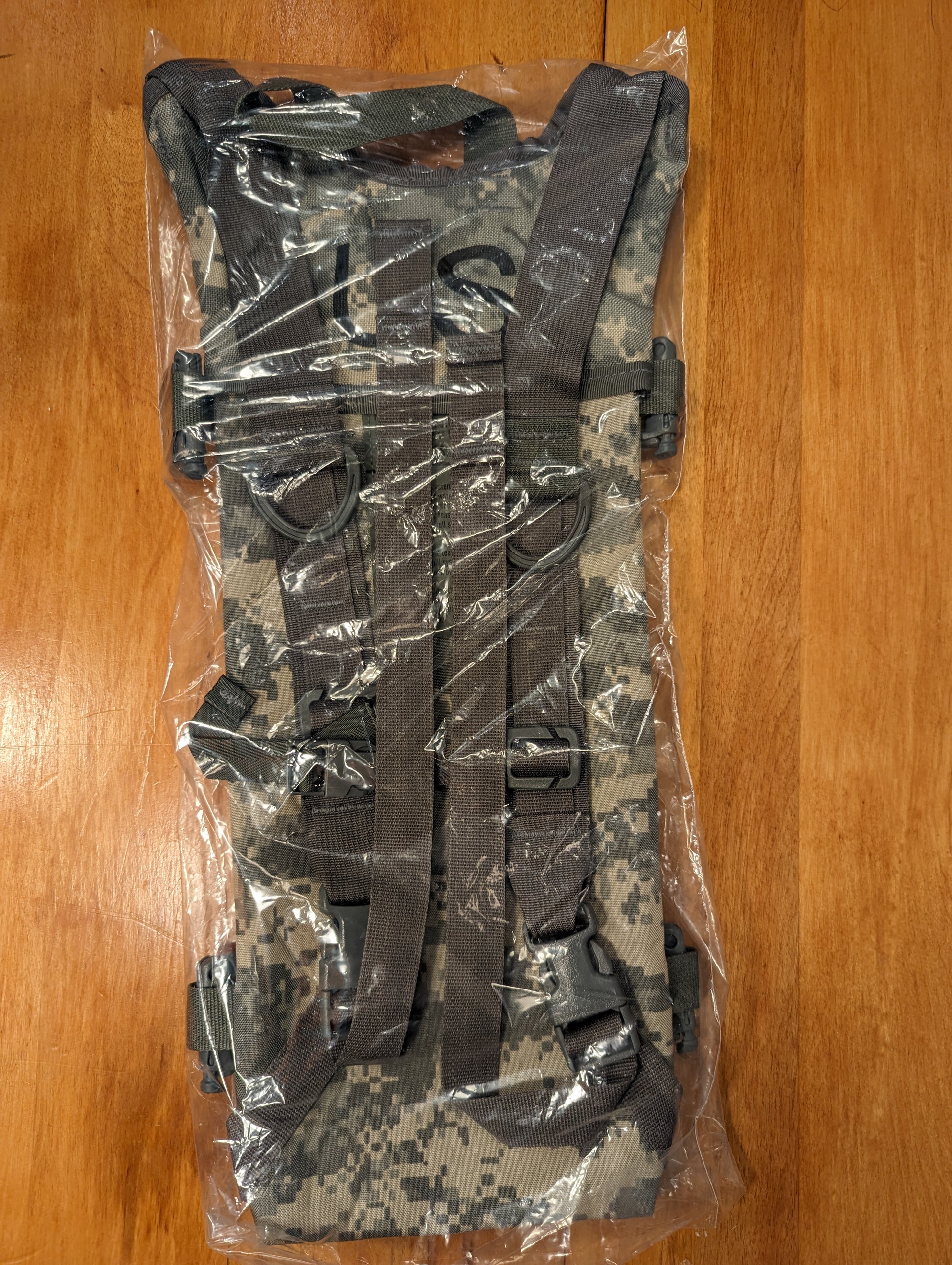 A new U.S. Military Hydration Carrier in ACU/UCP digital camouflage, sealed inside clear plastic packaging, showing the complete shoulder harness, chest strap, and black quick-release buckles.
