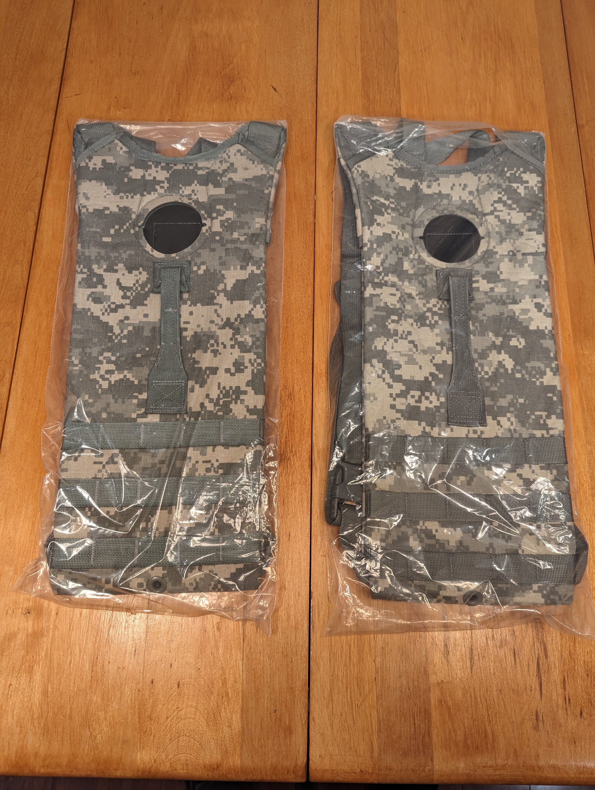 Two new U.S. Military Surplus Hydration Carriers in ACU/UCP digital camouflage, sealed inside clear plastic packaging, showing the pattern and top filler port on a wooden background.