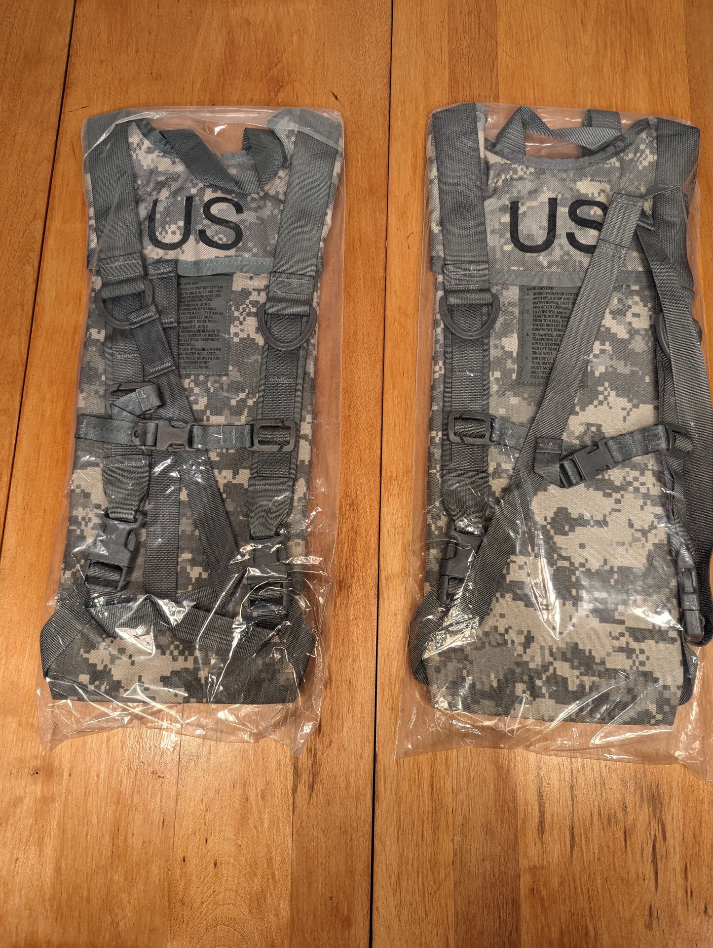Two new U.S. Military Surplus Hydration Carriers in ACU/UCP digital camouflage, sealed inside clear plastic packaging, featuring the large stenciled 'US' mark on the front of each carrier.