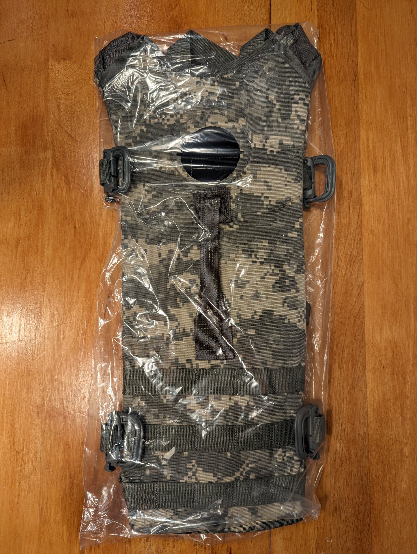 A new U.S. Military Hydration Carrier in ACU/UCP digital camouflage, sealed inside clear plastic packaging, showcasing the external MOLLE webbing, black buckles, and the circular filler port in the center.