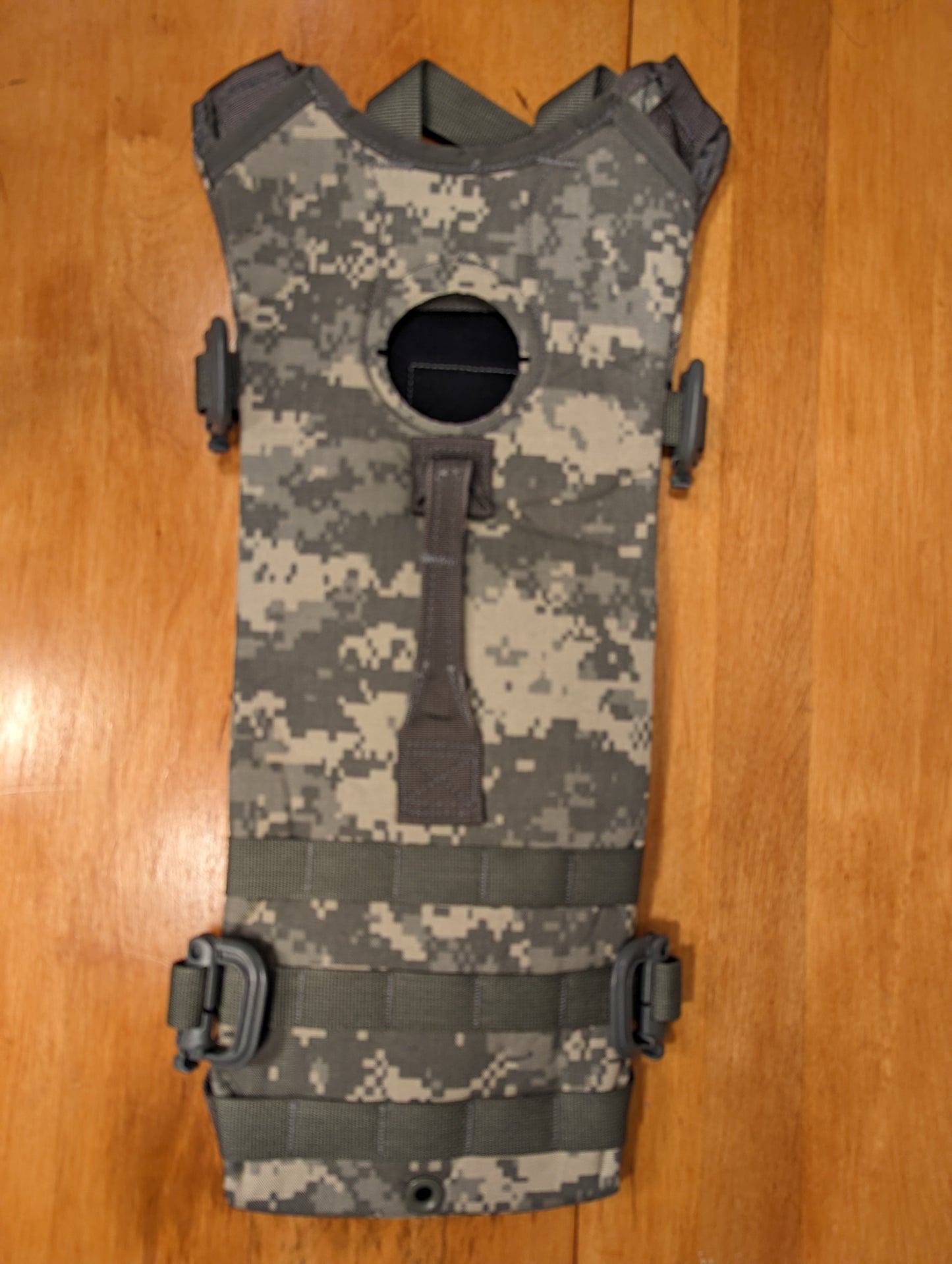 A U.S. Military Surplus Hydration Carrier pack shell in ACU digital camouflage, laid flat on a wooden surface, showcasing the MOLLE webbing and the rigid, structured profile.