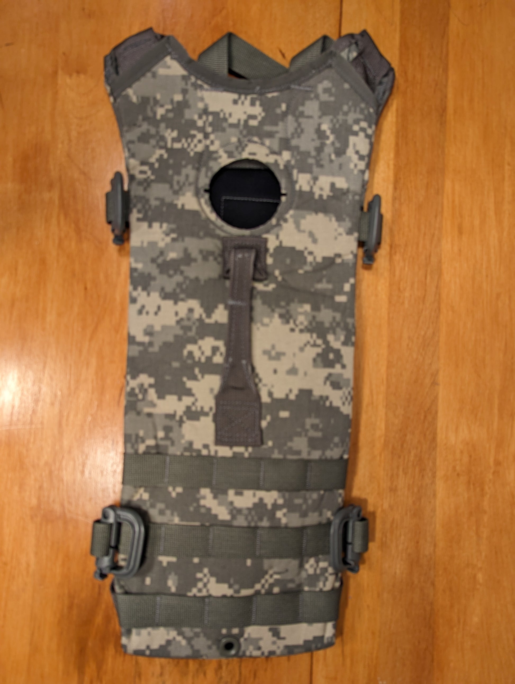 A U.S. Military Surplus Hydration Carrier pack shell in ACU digital camouflage, laid flat on a wooden surface, showcasing the MOLLE webbing and the rigid, structured profile.