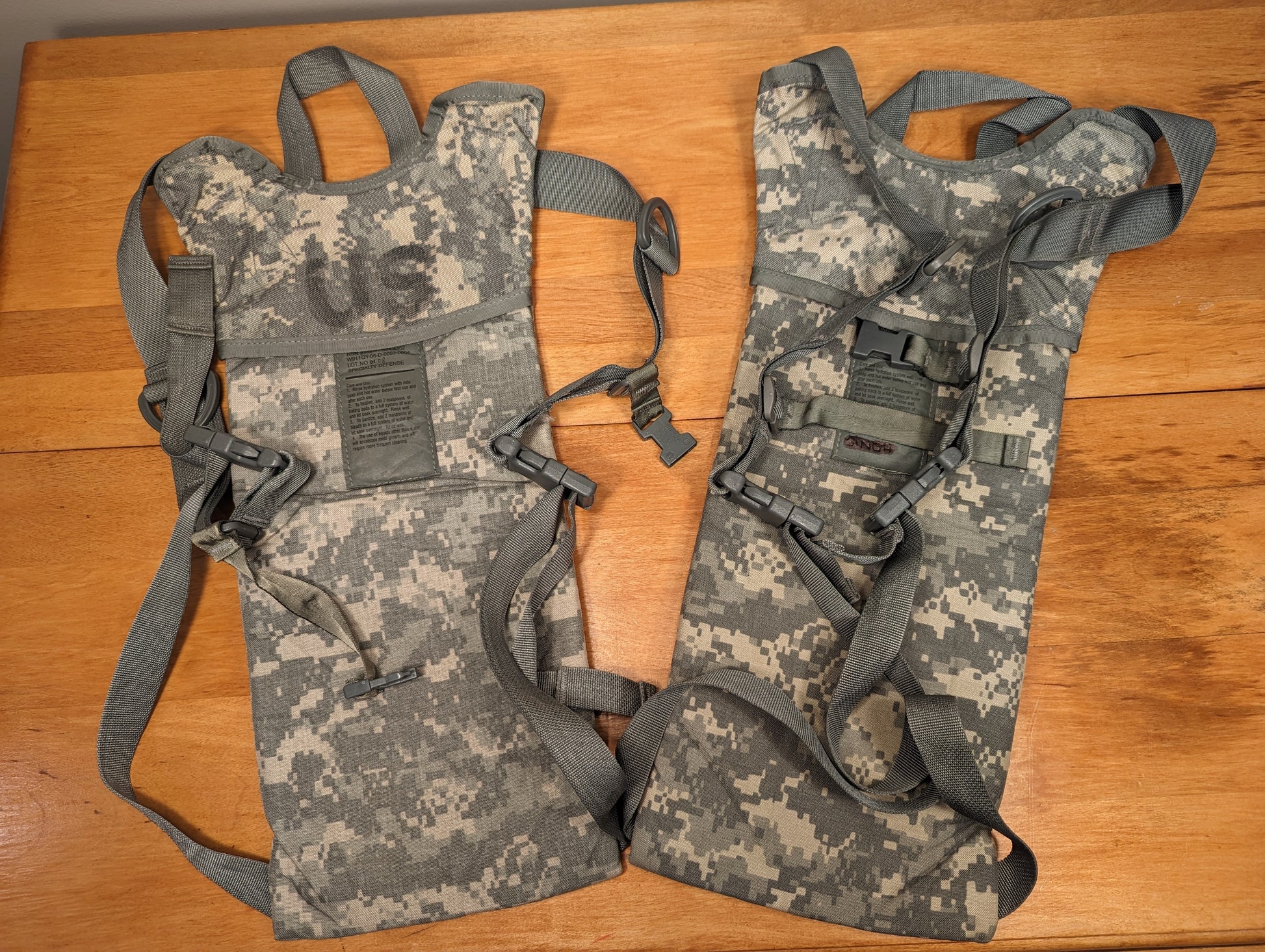 A pair of used U.S. Military Hydration Carriers in ACU digital camouflage, laid flat on a wooden table, showing the full padded shoulder harnesses and visible signs of field wear.
