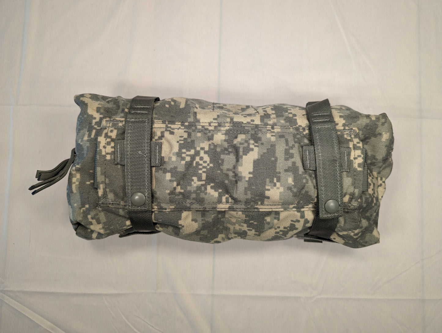 A tightly rolled U.S. Military Surplus Waist Pack in ACU digital camouflage, secured by two black webbing straps with snap closures, resting horizontally on a white surface.