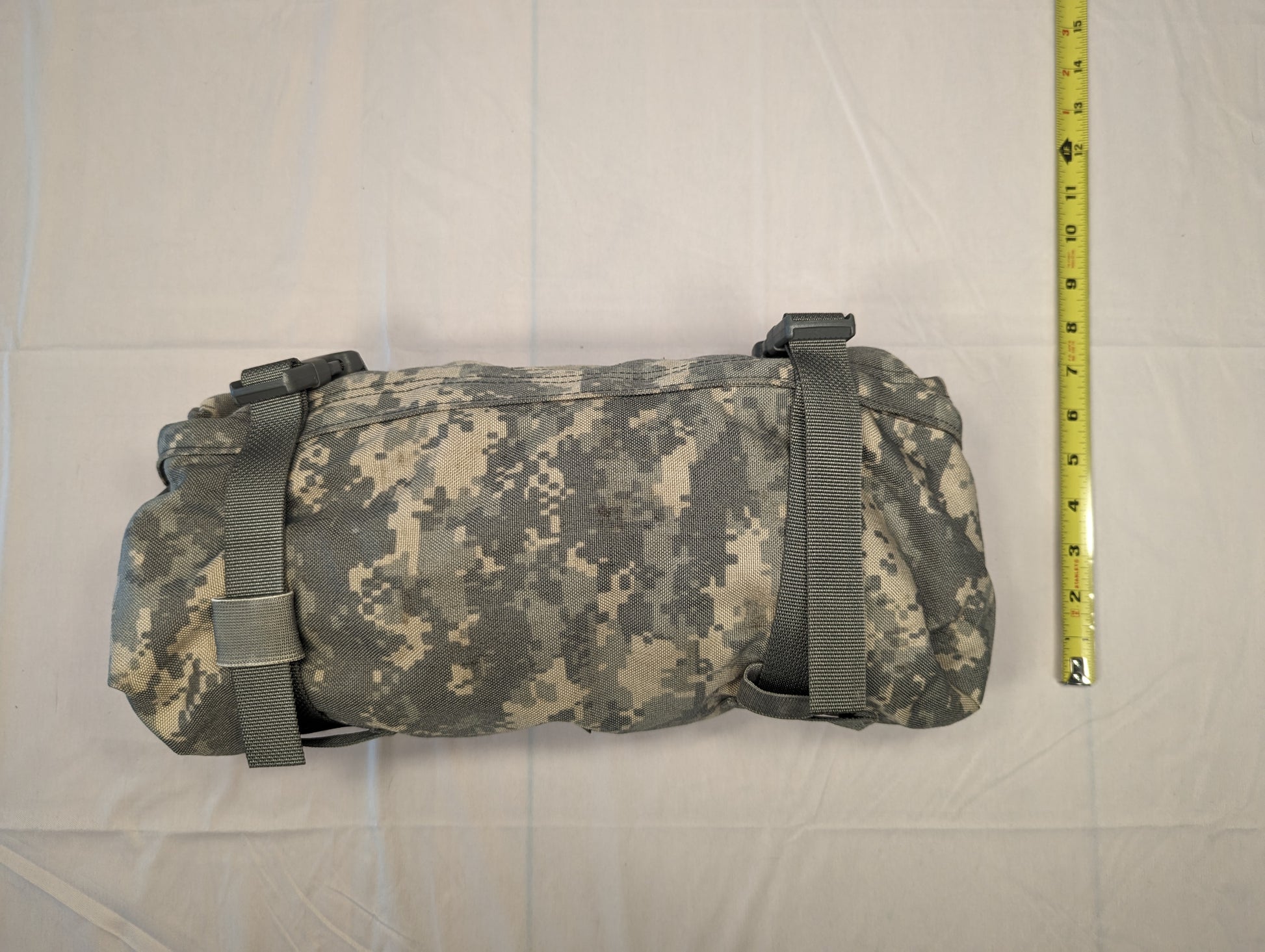 A U.S. Military Surplus Waist Pack in ACU digital camouflage, tightly rolled and secured by webbing straps, with a yellow tape measure placed alongside to demonstrate its compact size.