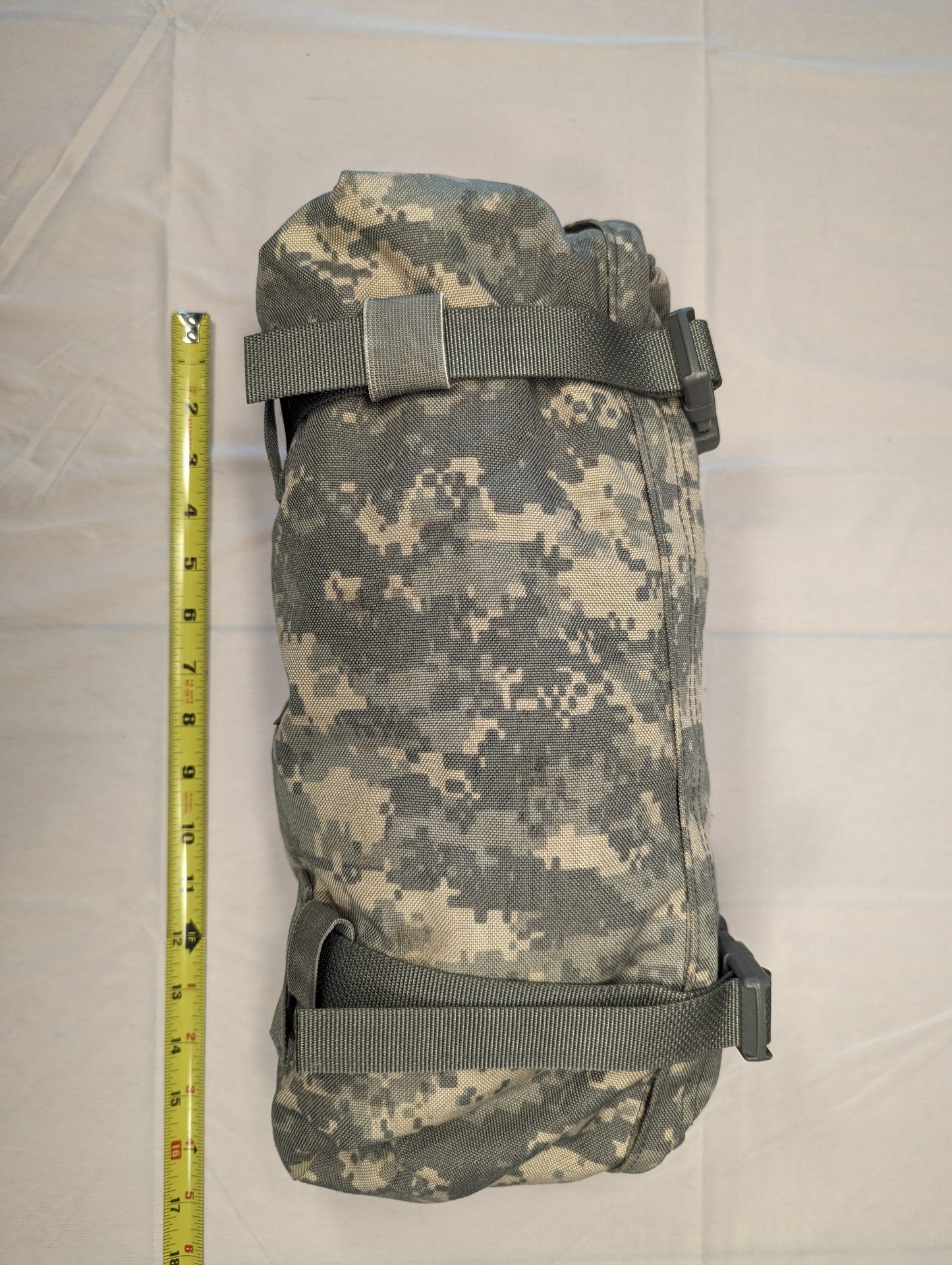 A U.S. Military Surplus Waist Pack in ACU digital camouflage, tightly rolled and secured by webbing straps, with a yellow tape measure placed alongside to demonstrate its compressed size of approximately 14 inches.