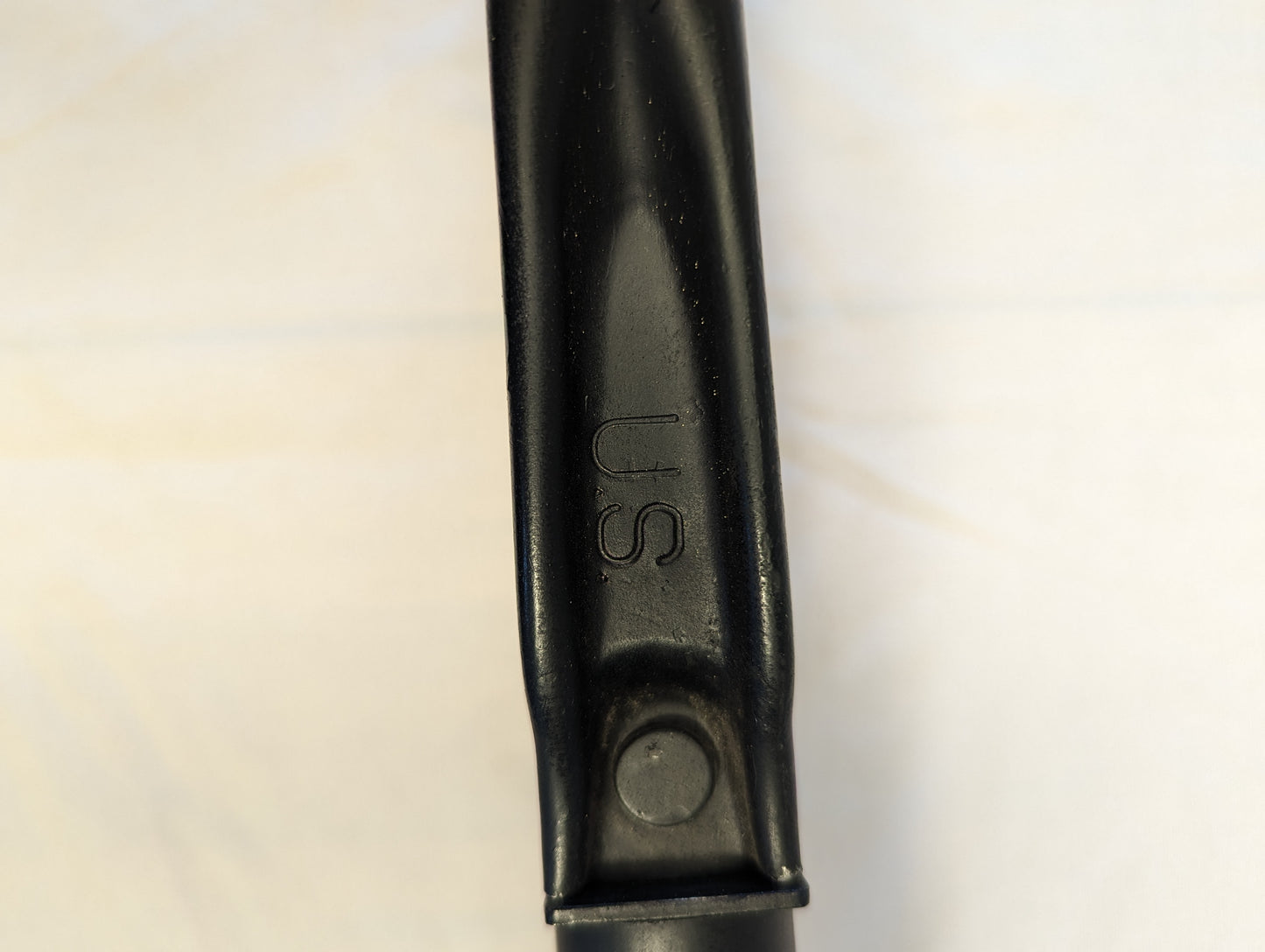 A close-up photograph of the black metal handle of the U.S. Military AMES Entrenching Tool, showing the embossed 'U.S.' government property marking and an attachment rivet.