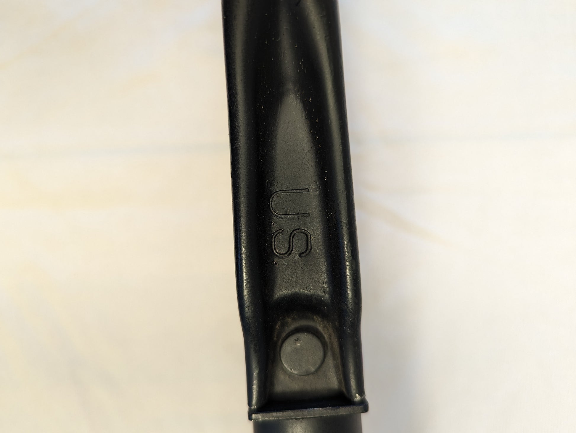 A close-up photograph of the black metal handle of the U.S. Military AMES Entrenching Tool, showing the embossed 'U.S.' government property marking and an attachment rivet.