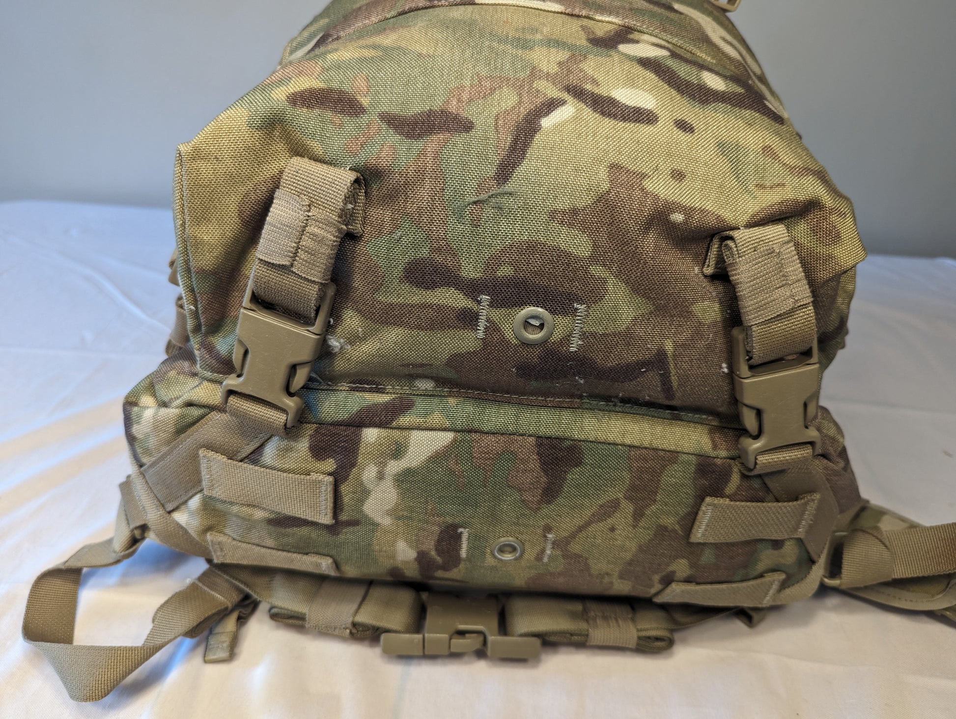A close-up view of the bottom panel of a U.S. Military Assault Pack in OCP/Multicam camouflage, highlighting the MOLLE webbing, tan quick-release buckles, and metal drainage grommets.