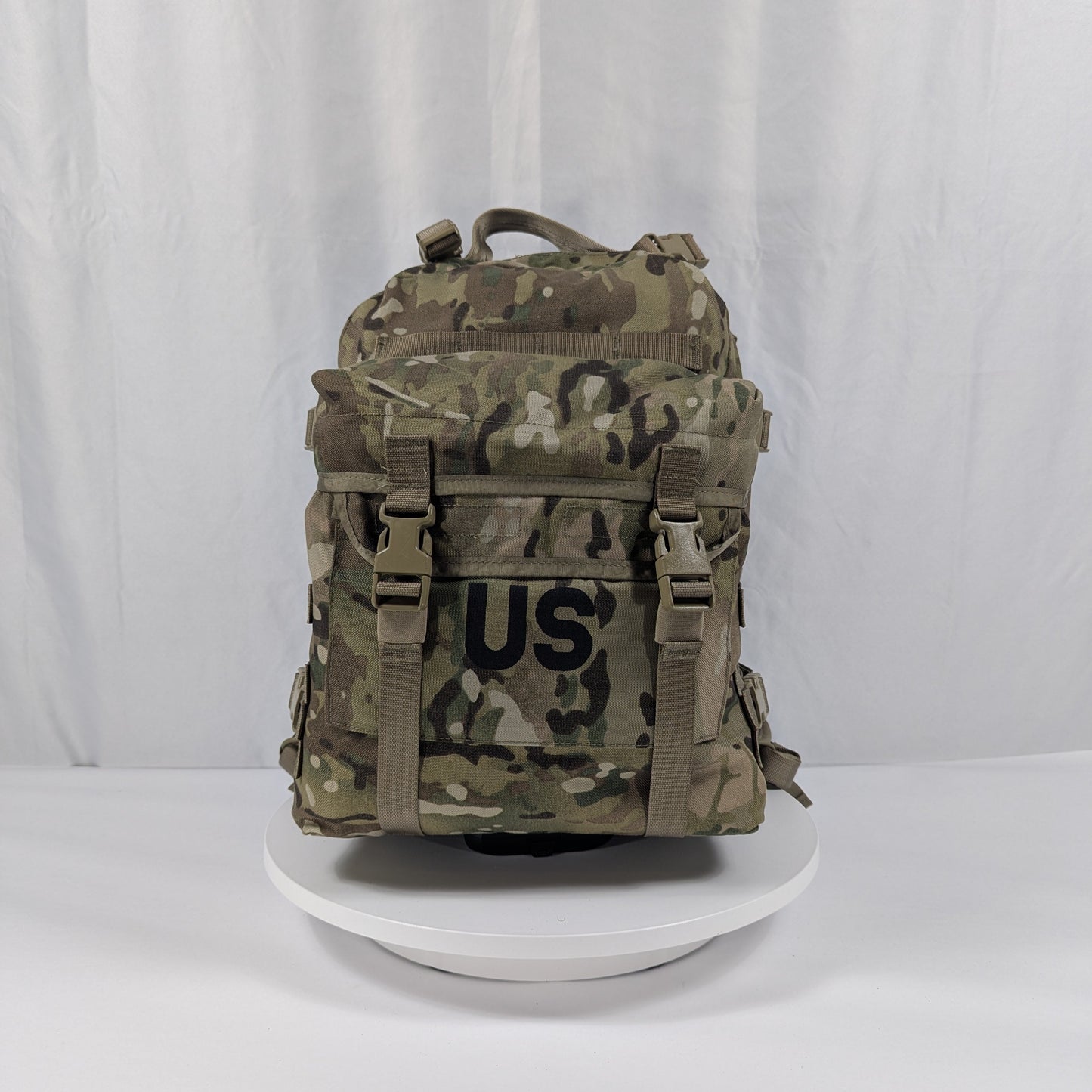 A U.S. Military Surplus 3-Day Assault Pack in OCP/Multicam camouflage, standing upright on a white display, prominently featuring a large stenciled 'US' mark, horizontal MOLLE webbing, and adjustable compression straps.