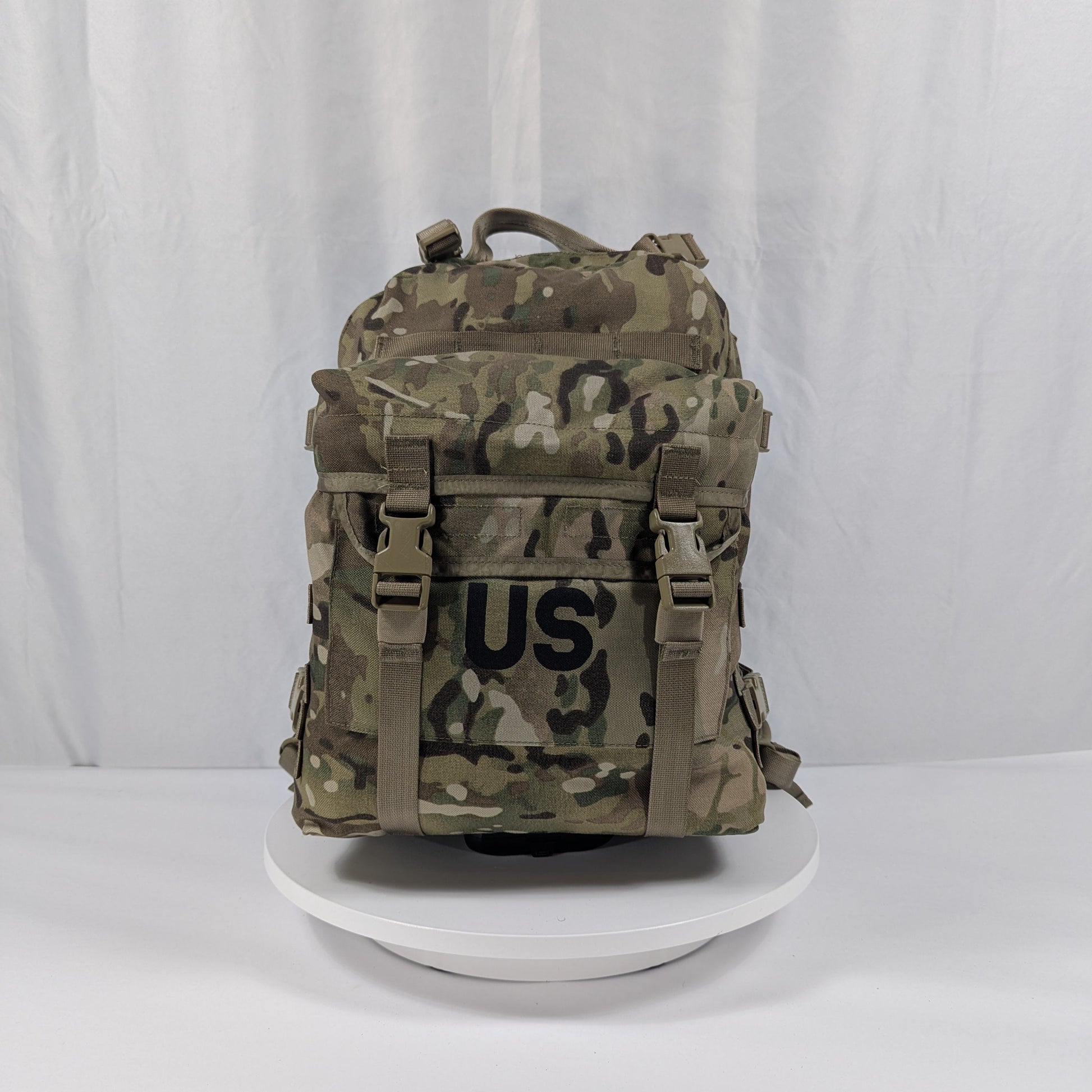 A U.S. Military Surplus 3-Day Assault Pack in OCP/Multicam camouflage, standing upright on a white display, prominently featuring a large stenciled 'US' mark, horizontal MOLLE webbing, and adjustable compression straps.