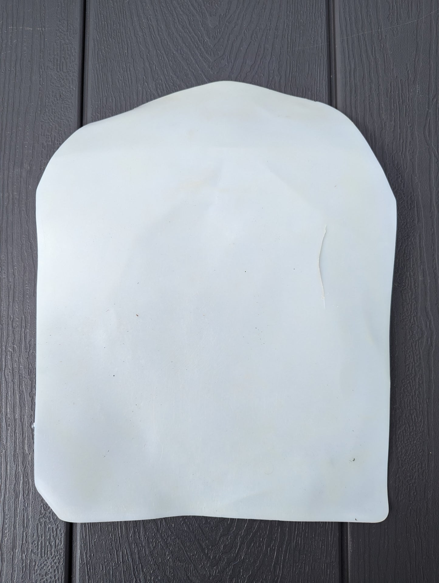 A single, white, semi-rigid plastic stiffener pad from the U.S. Military Surplus Assault Pack, resting on a dark, ridged wooden surface.