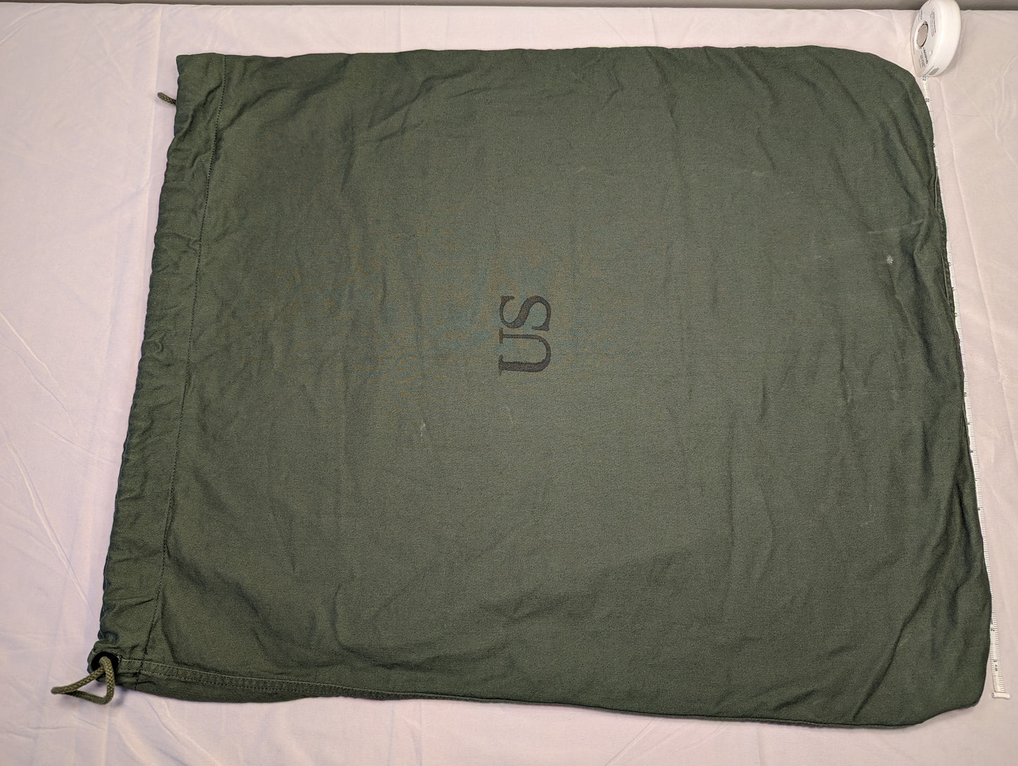 A large, olive green U.S. Military Surplus Barracks Bag laid flat, prominently featuring a stenciled 'US' mark in the center and a drawstring closure at the corner.