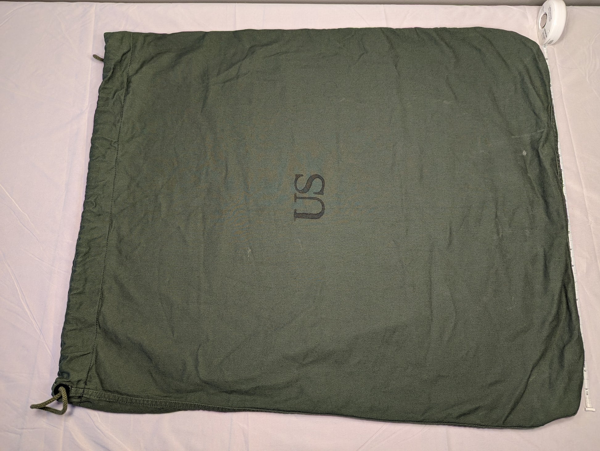 A large, olive green U.S. Military Surplus Barracks Bag laid flat, prominently featuring a stenciled 'US' mark in the center and a drawstring closure at the corner.