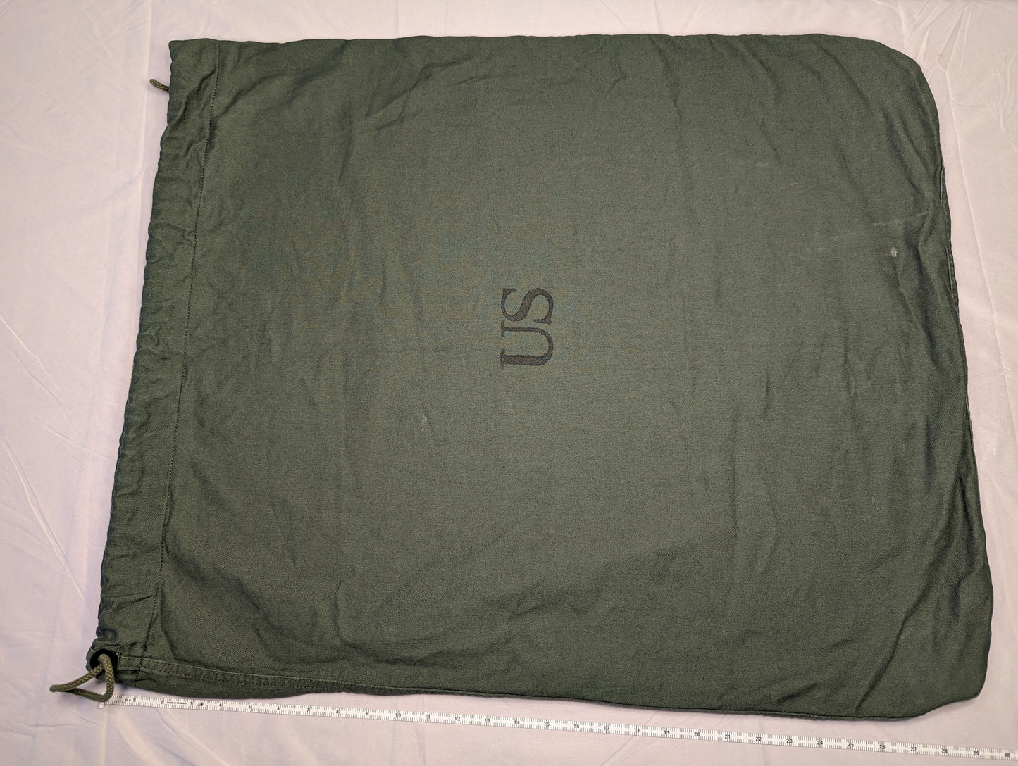 A large, olive green U.S. Military Surplus Barracks Bag laid flat, prominently featuring a stenciled 'US' mark, a drawstring closure, and a measuring tape along the bottom edge demonstrating its width.
