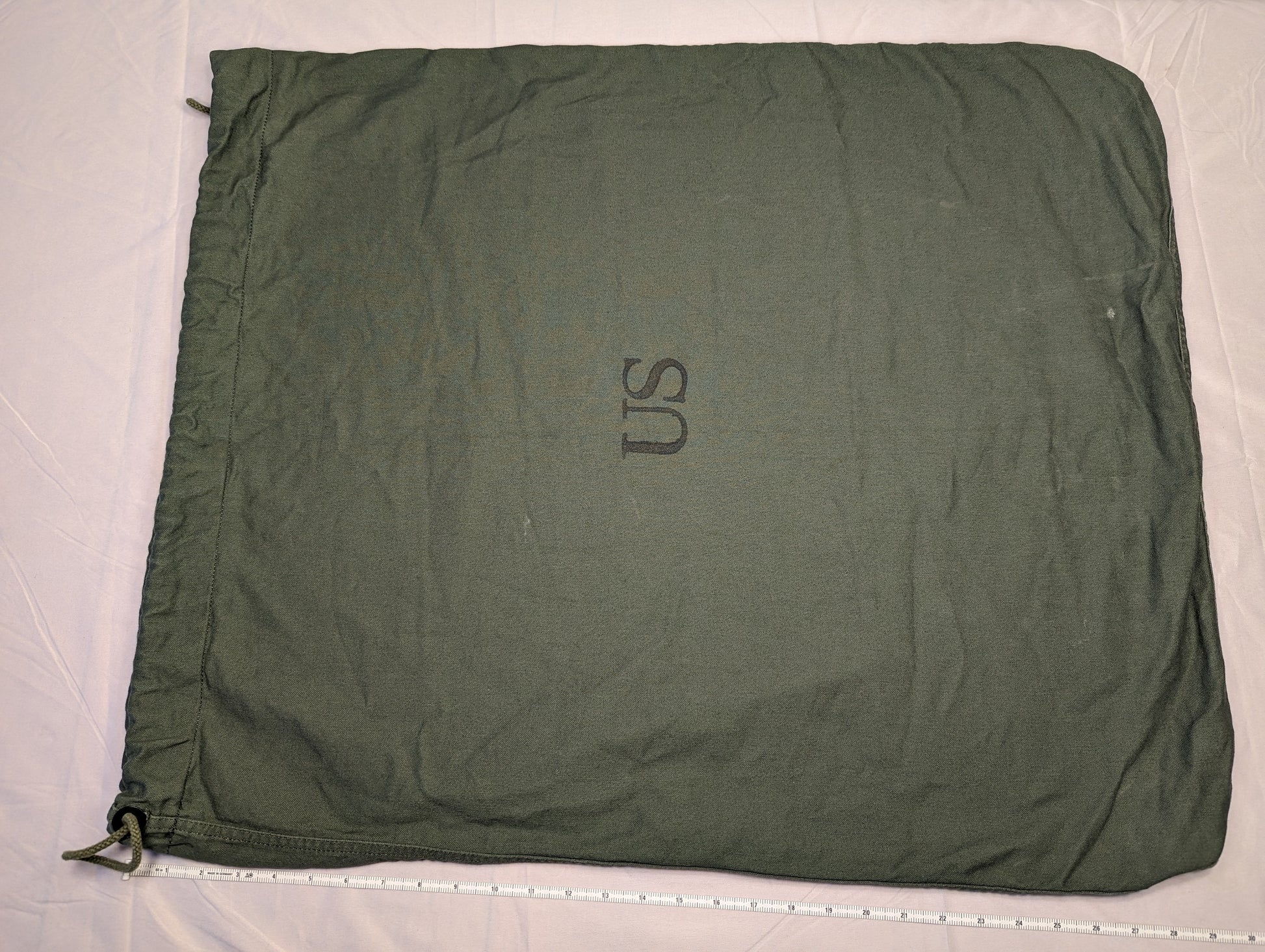 A large, olive green U.S. Military Surplus Barracks Bag laid flat, prominently featuring a stenciled 'US' mark, a drawstring closure, and a measuring tape along the bottom edge demonstrating its width.