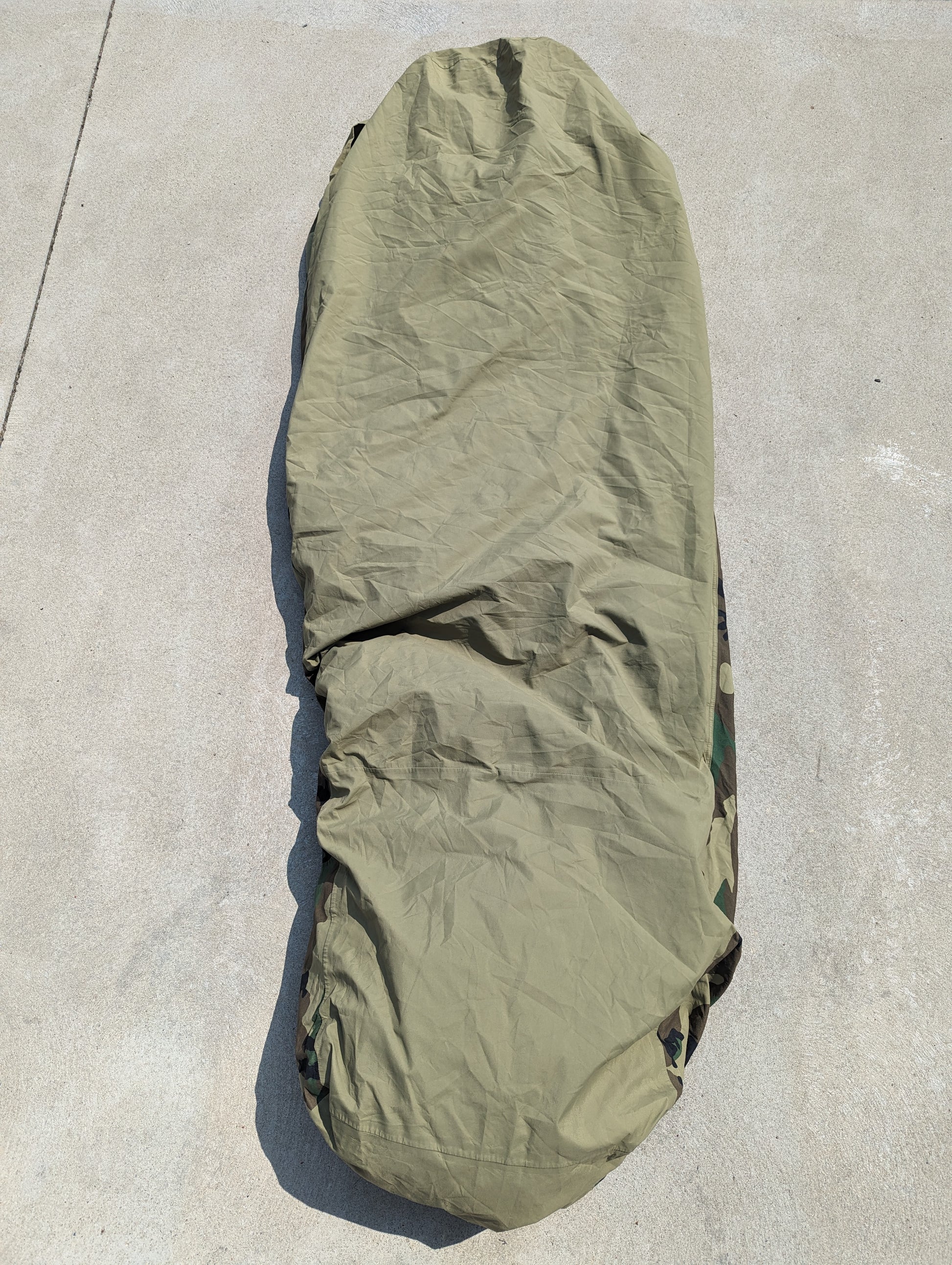 A full-length U.S. Military Surplus Modular Sleep System Bivy Cover in solid olive green, zipped closed and laid flat on a concrete surface, displaying its wrinkled, waterproof fabric.