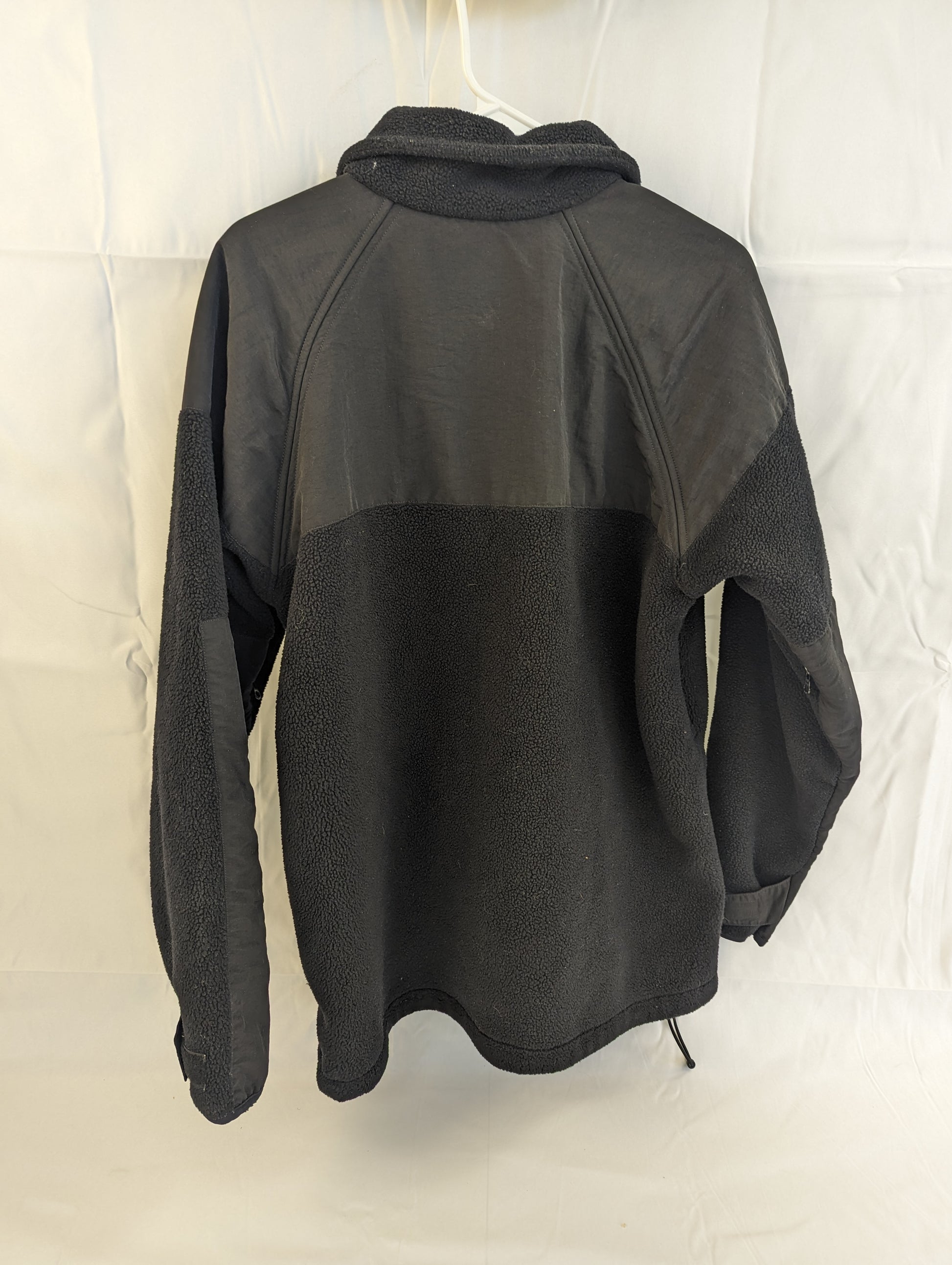 A back view of a black U.S. Military Cold Weather Fleece Jacket hanging on a hanger, highlighting the reinforced nylon panels on the shoulders and upper back.