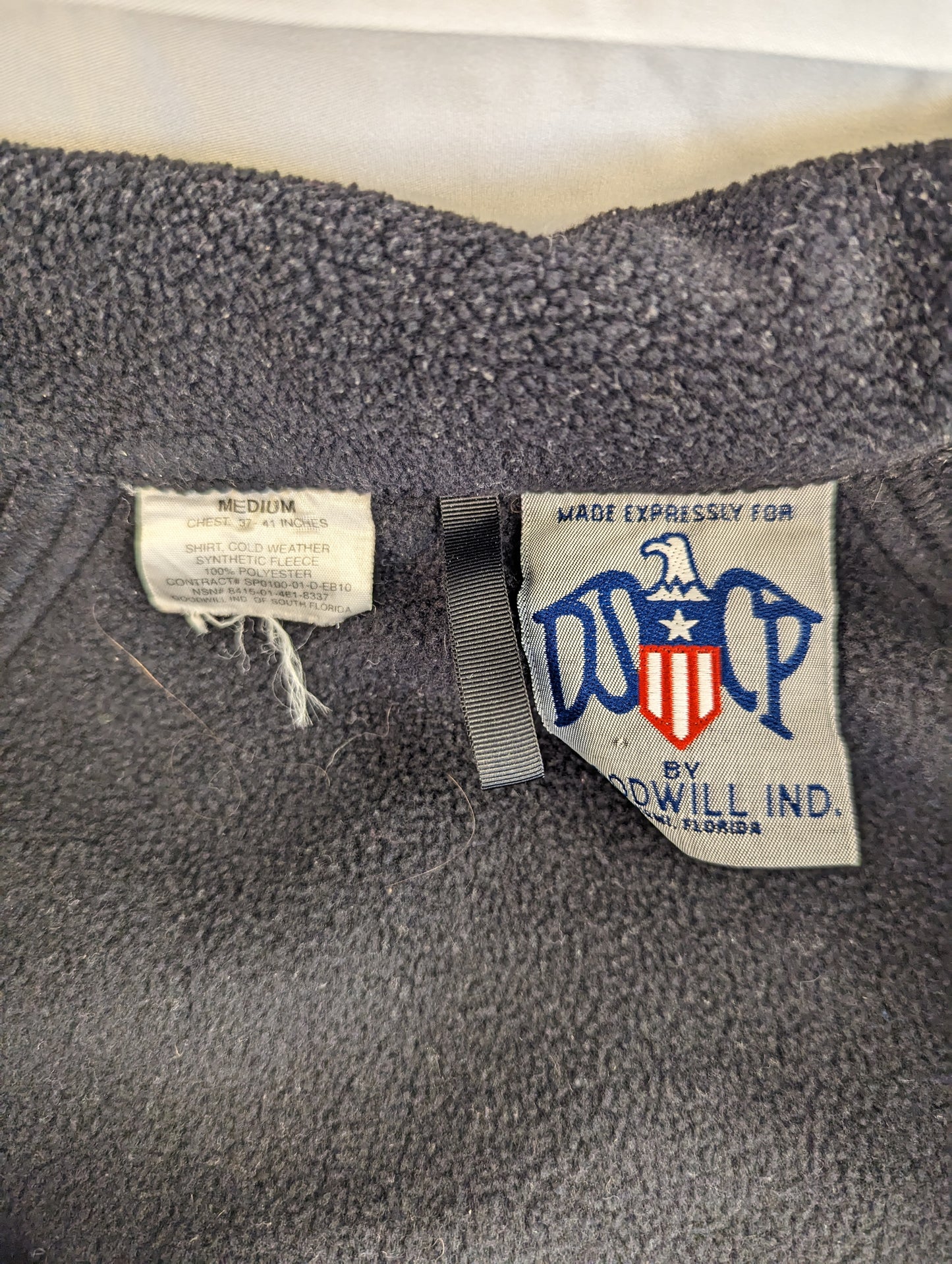 A close-up of the interior neck tags of a black U.S. Military Cold Weather Fleece Jacket, displaying the 'Made Expressly for DSCP by Goodwill Ind. of South Florida' logo and the size 'MEDIUM' label.
