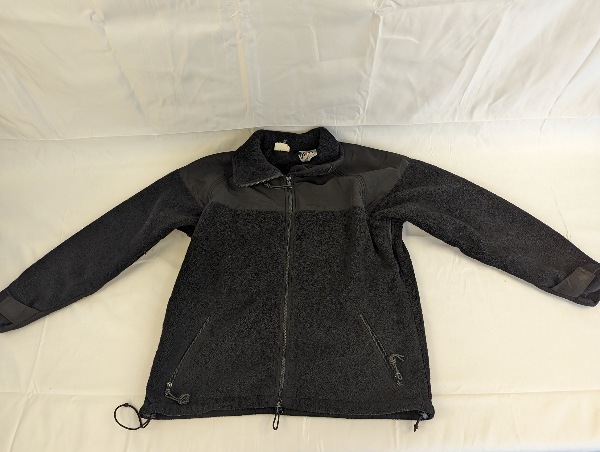 A front view of a black U.S. Military Cold Weather Fleece Jacket laid flat on a white background, featuring reinforced nylon shoulder and chest panels, a full-length zipper, and two lower pockets.