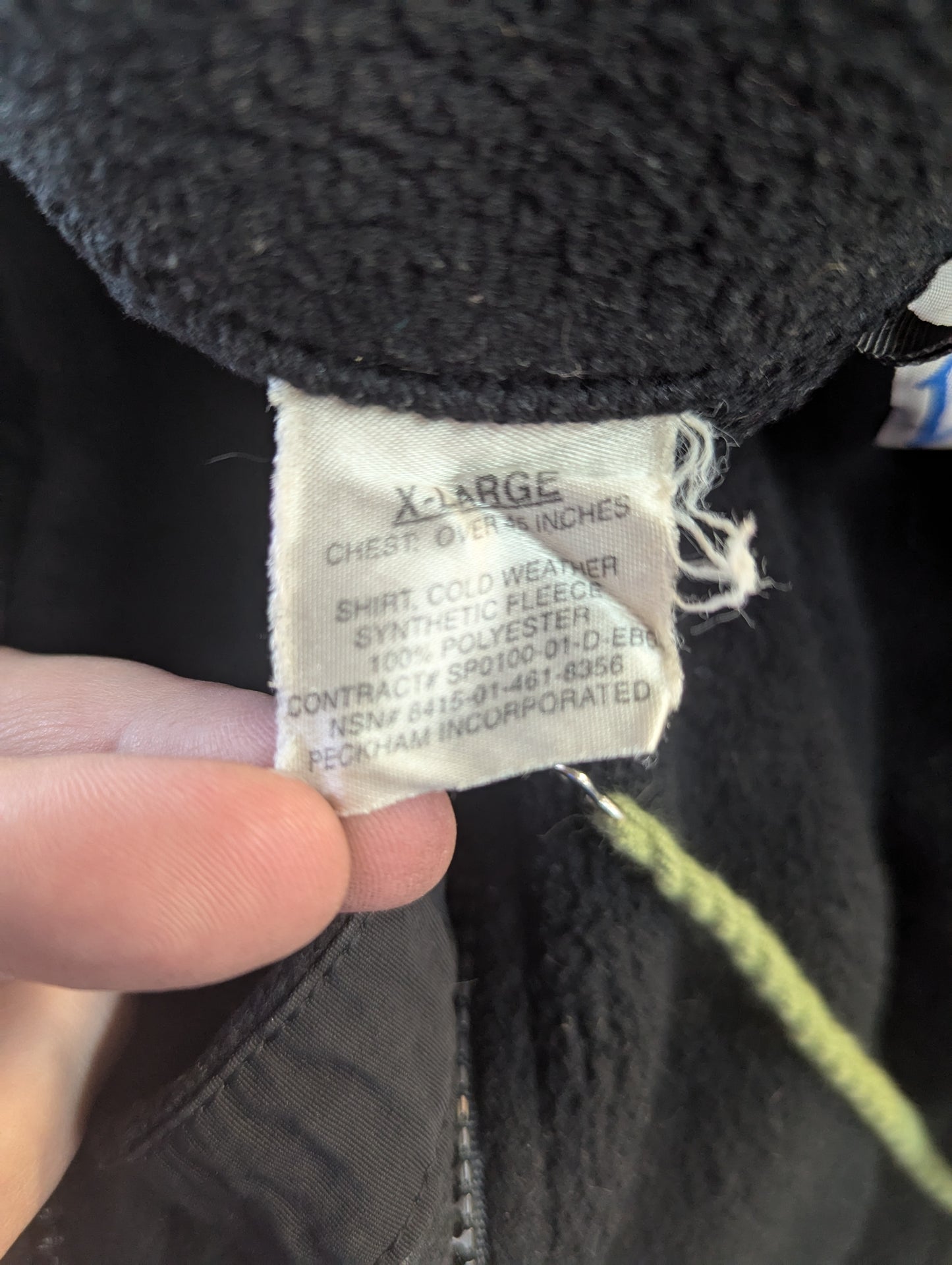 A close-up photograph of the U.S. Military issue tag sewn into the black cold weather fleece, displaying the size 'X-LARGE', the National Stock Number (NSN) 8415-01-461-8356, and manufacturer Peckham Incorporated.