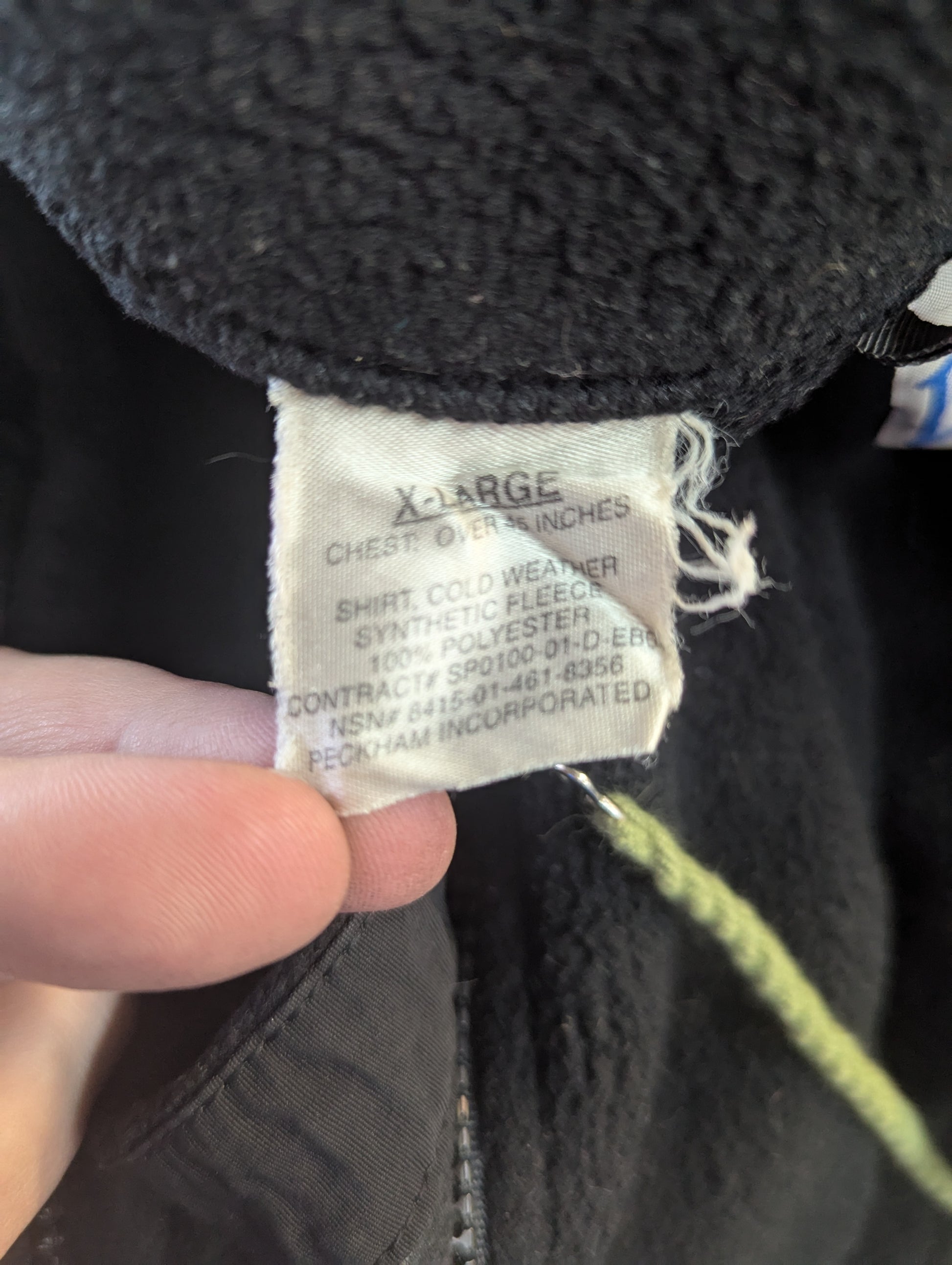 A close-up photograph of the U.S. Military issue tag sewn into the black cold weather fleece, displaying the size 'X-LARGE', the National Stock Number (NSN) 8415-01-461-8356, and manufacturer Peckham Incorporated.