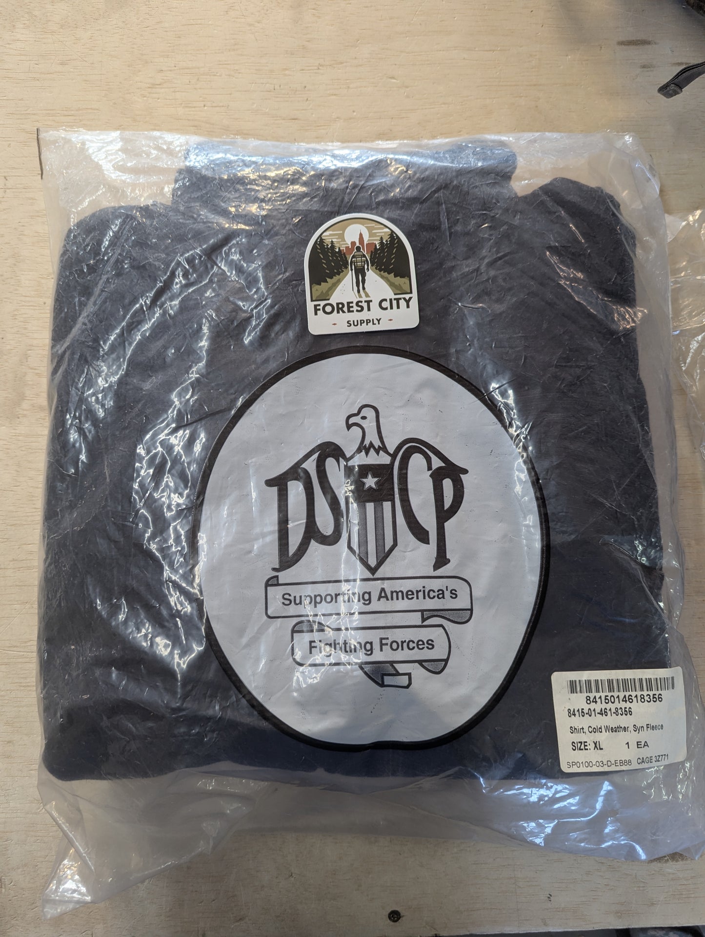 A brand new, packaged U.S. Military Cold Weather Fleece Jacket in black, sealed in a clear plastic bag featuring the large 'DSCP Supporting America's Fighting Forces' logo and a Forest City Supply sticker.