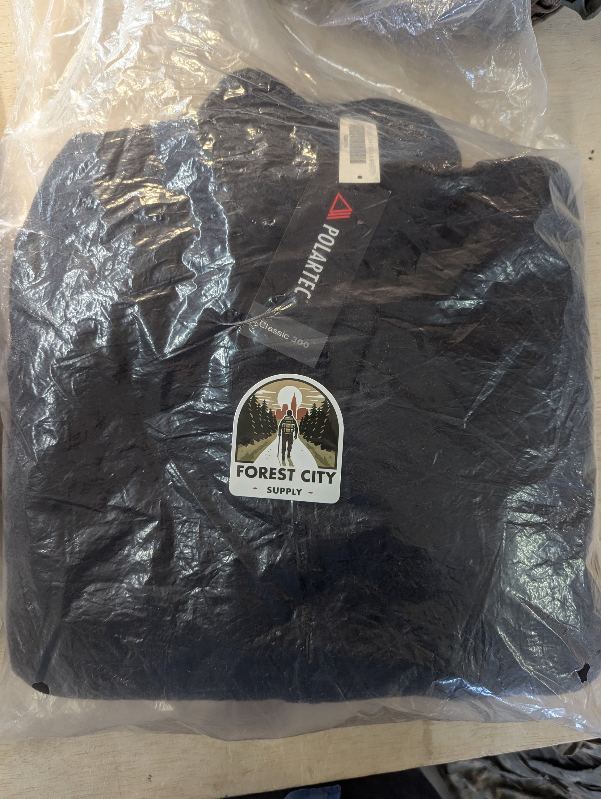 A brand new, packaged black Polartec fleece jacket sealed in clear plastic, featuring a 'FOREST CITY SUPPLY' logo sticker and visible manufacturer hang tags.