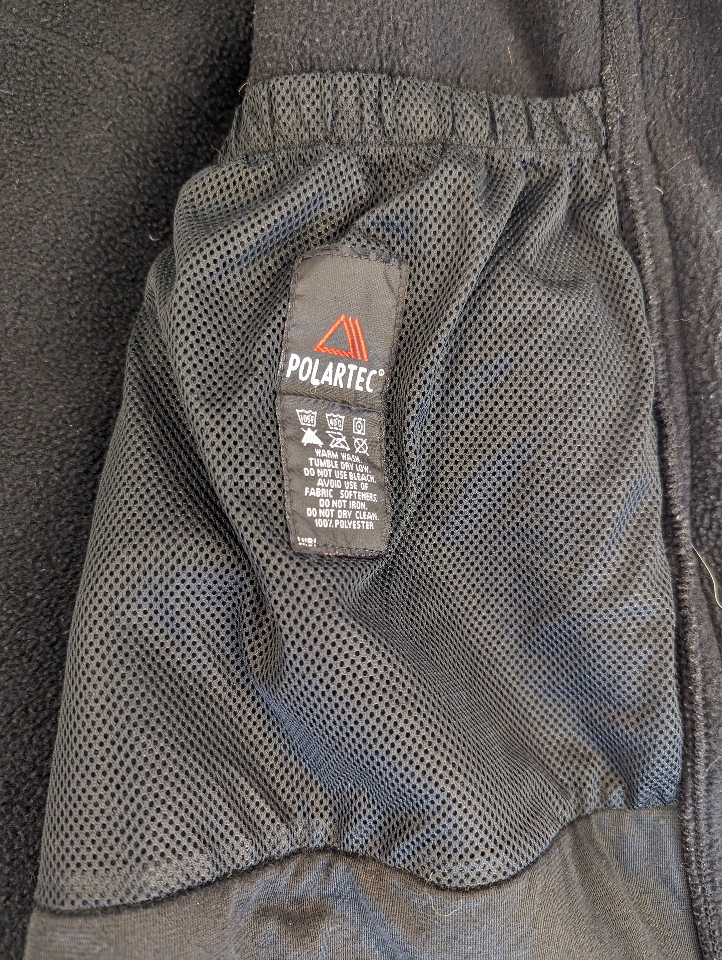 A close-up view of the interior mesh pocket of a black U.S. Military Cold Weather Fleece Jacket, clearly displaying the woven Polartec label with laundry symbols and care instructions.