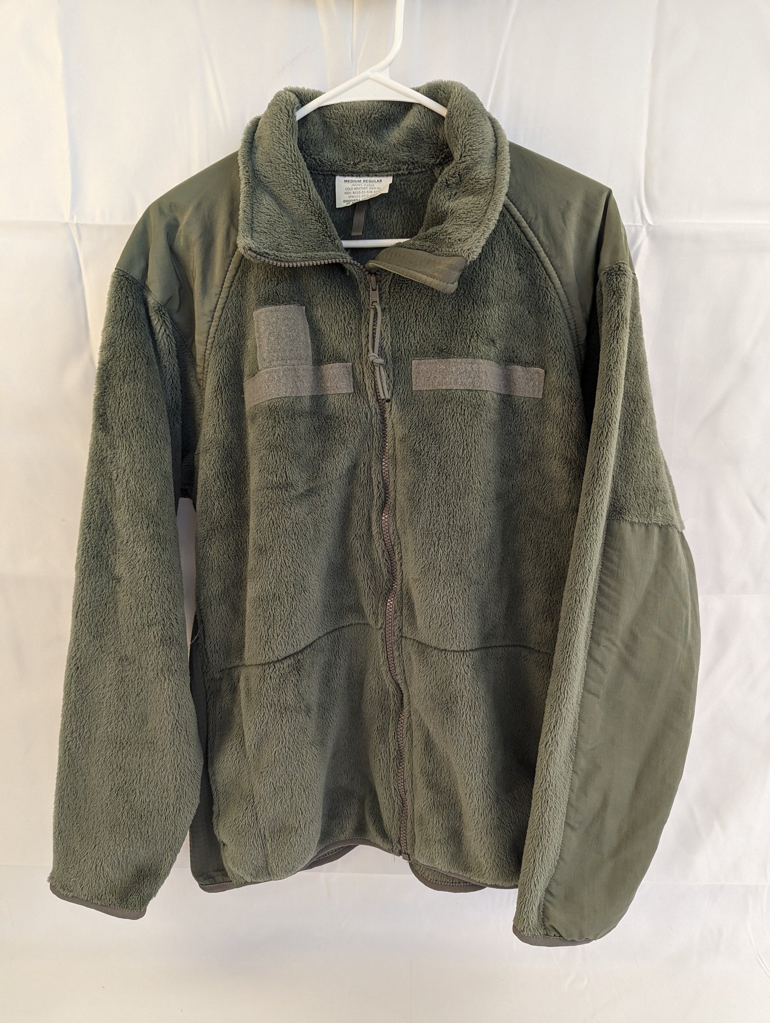 A front view of a U.S. Military Surplus Cold Weather Fleece Jacket in olive green, hanging on a white hanger, featuring reinforced nylon shoulder panels, a full-length zipper, and a high collar.