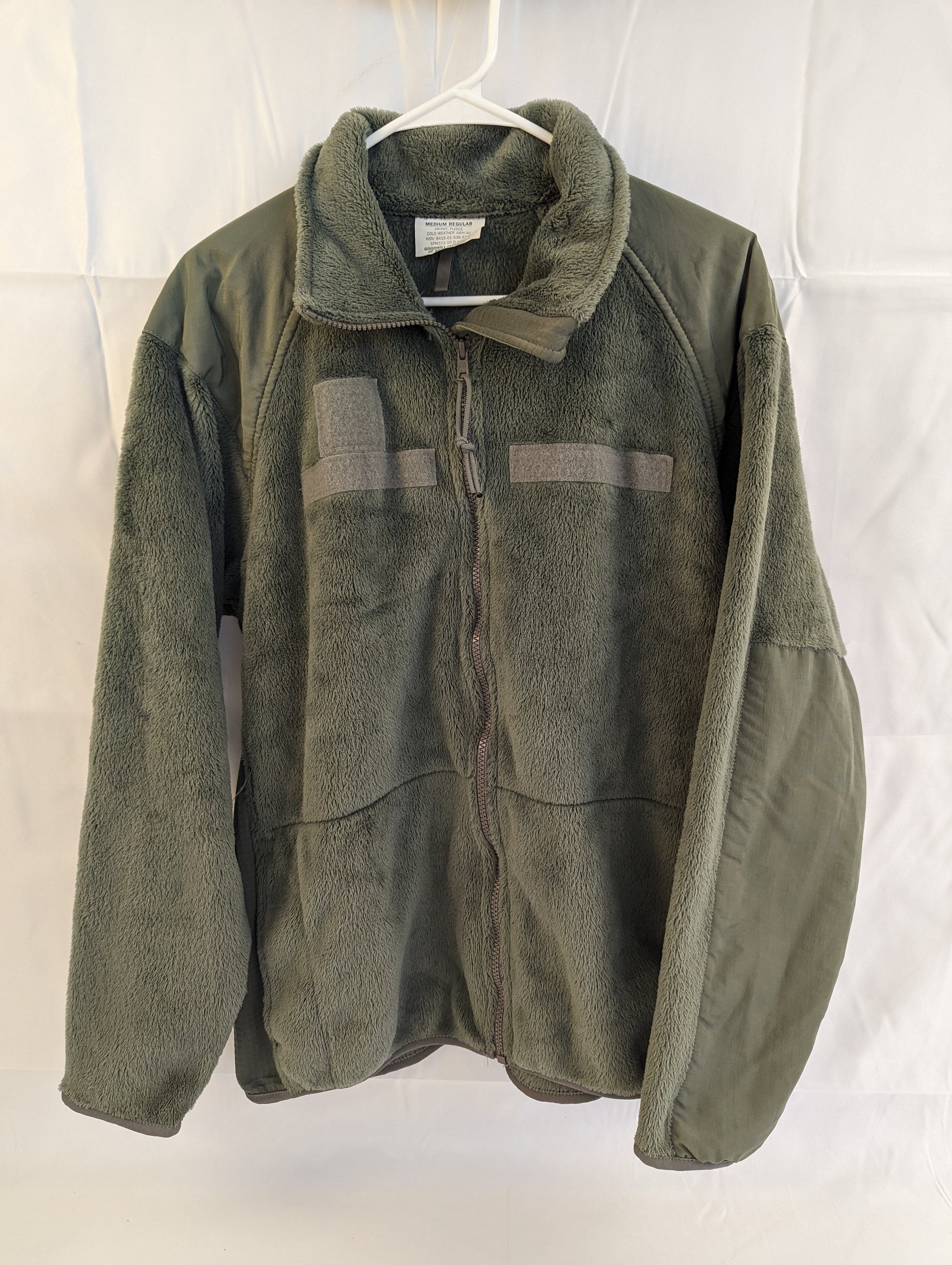 A front view of a U.S. Military Surplus Cold Weather Fleece Jacket in olive green, hanging on a white hanger, featuring reinforced nylon shoulder panels, a full-length zipper, and a high collar.