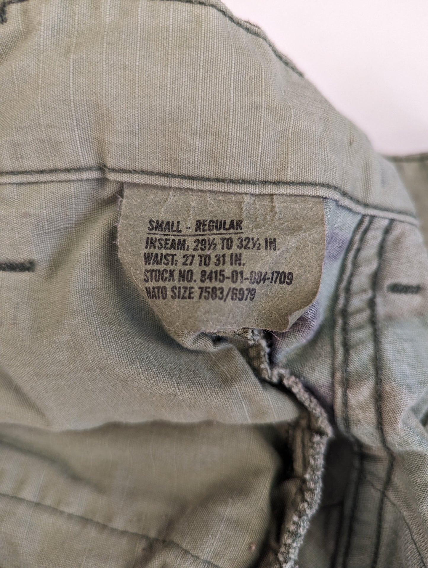 A close-up photograph of the U.S. Military issue sizing tag sewn into the combat trousers, clearly displaying the size 'SMALL-REGULAR', waist, inseam, and stock numbers.