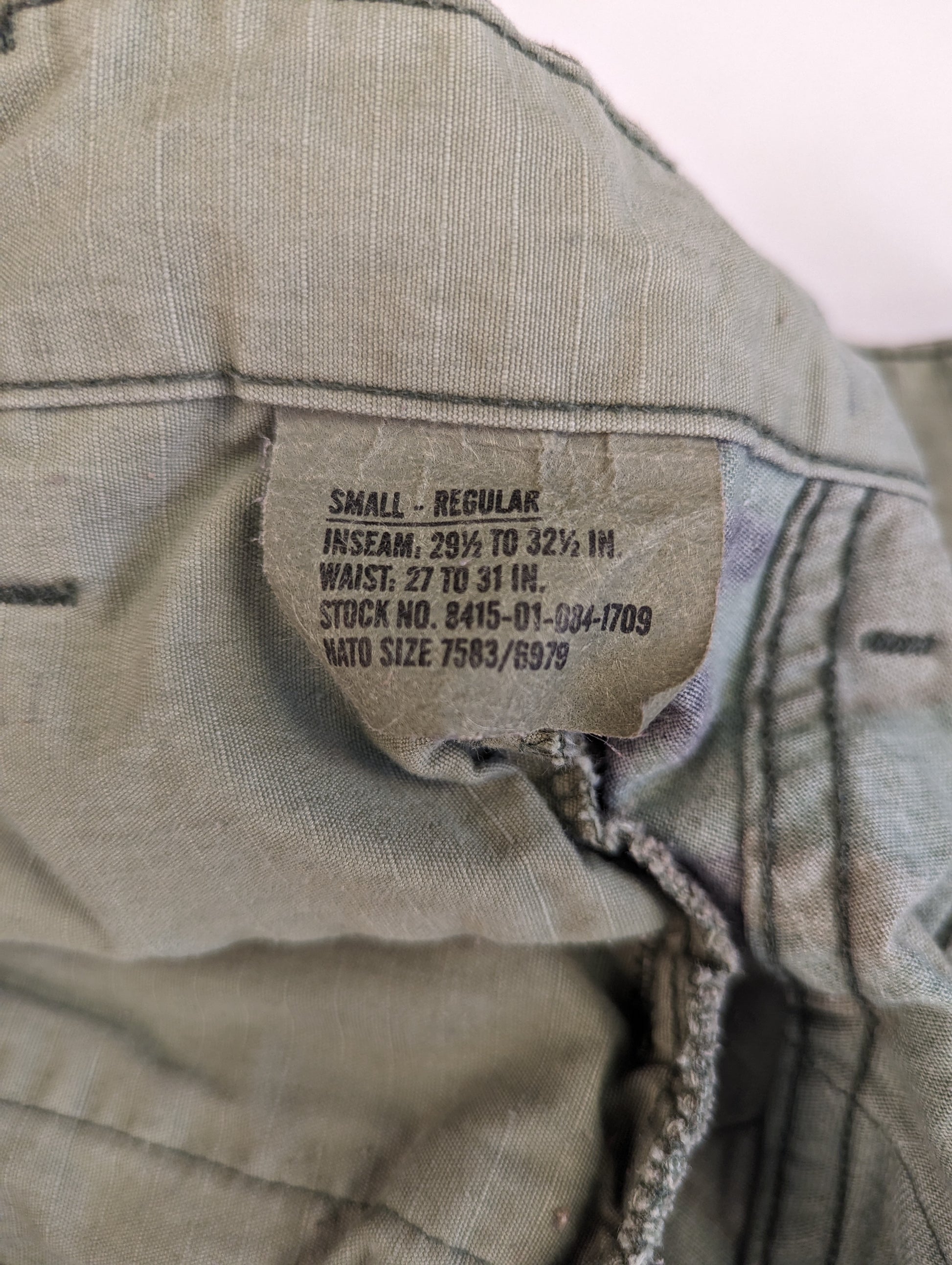 A close-up photograph of the U.S. Military issue sizing tag sewn into the combat trousers, clearly displaying the size 'SMALL-REGULAR', waist, inseam, and stock numbers.