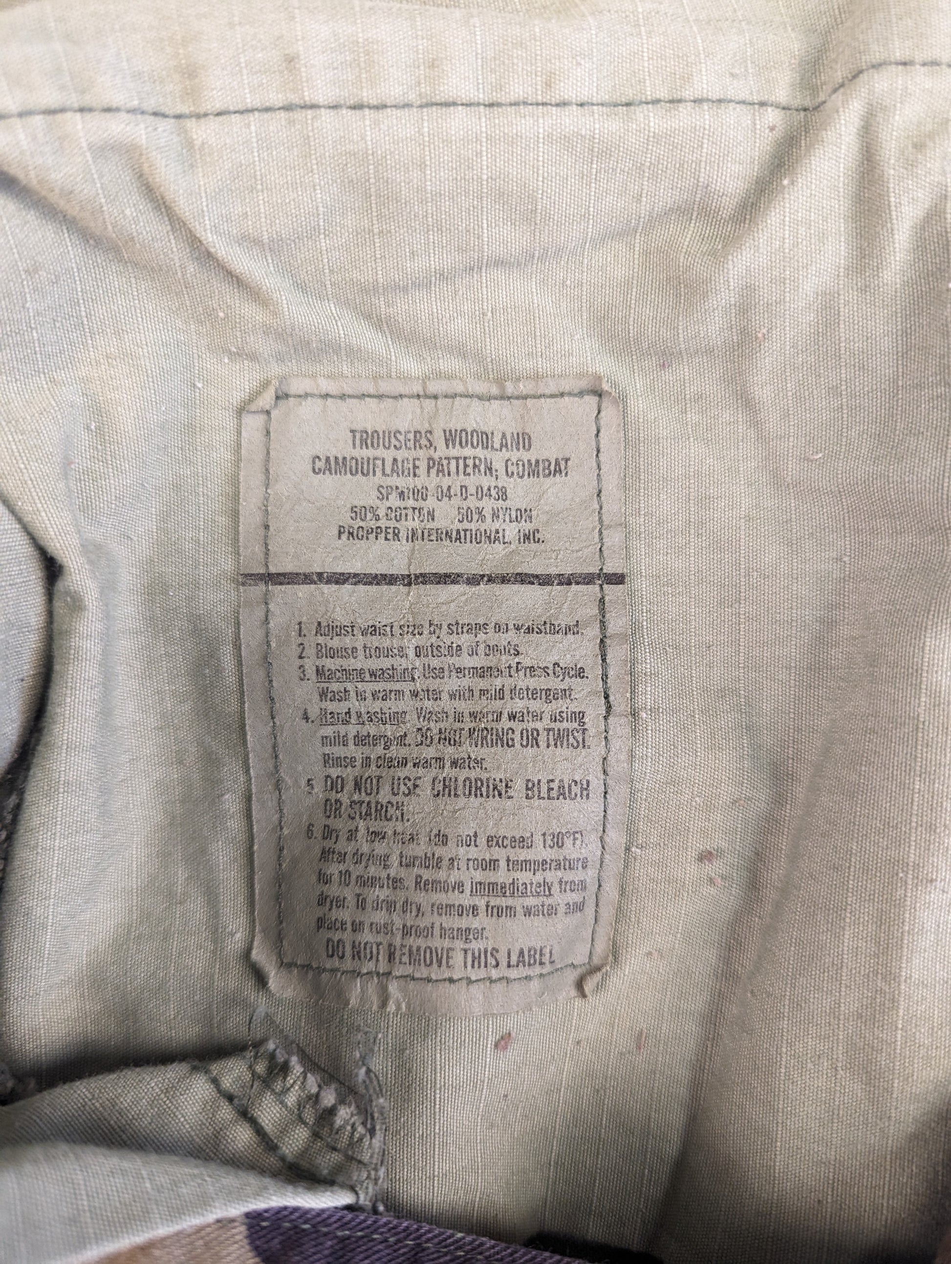 A close-up photograph of the original manufacturer's care tag sewn into the Woodland Camouflage Combat Trousers, displaying detailed washing and drying instructions.