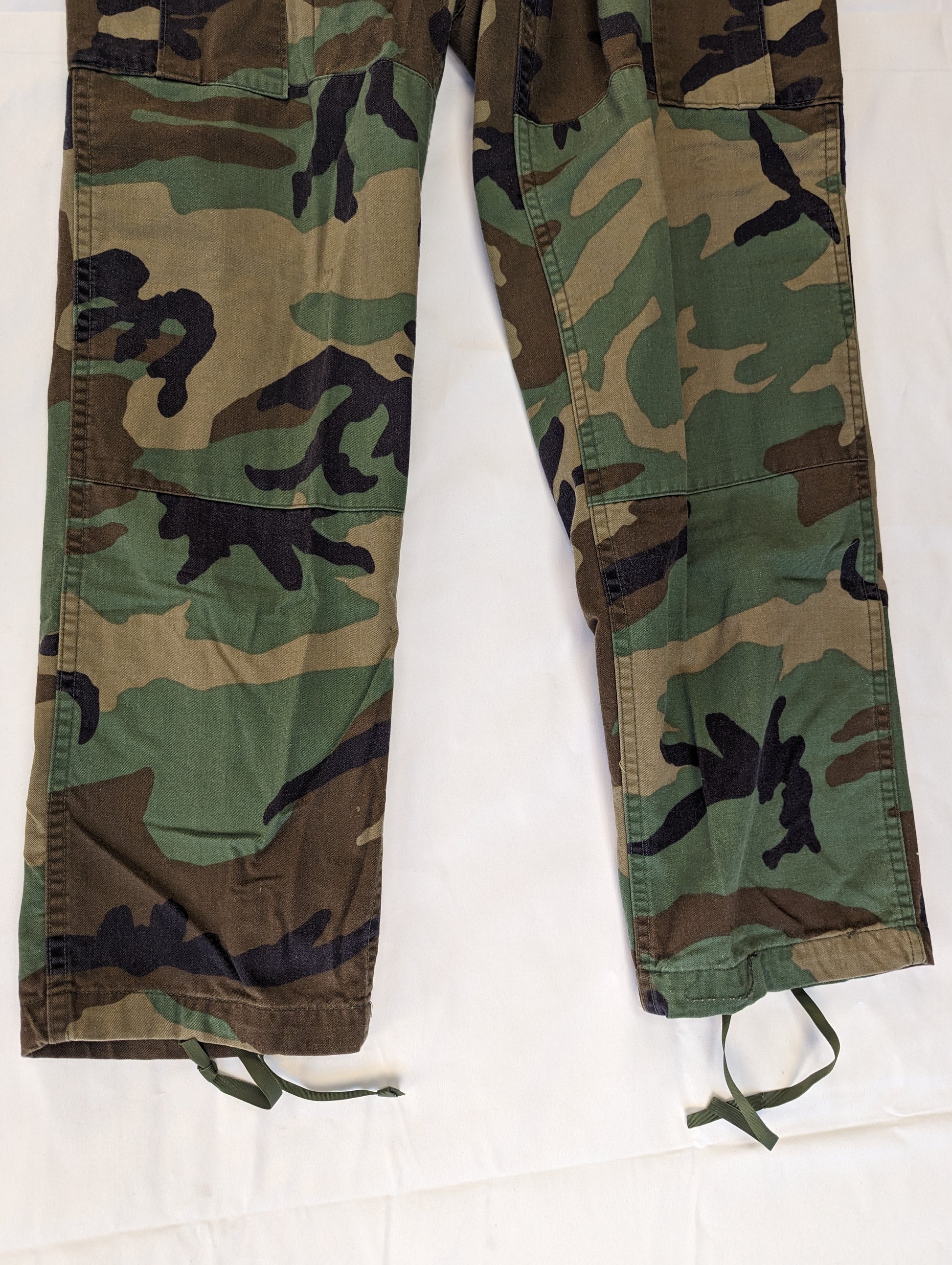 A close-up studio shot of the lower legs of a pair of U.S. Military Surplus Combat Trousers in Woodland camouflage, highlighting the texture of the fabric, knee reinforcement, and drawstring cuffs.