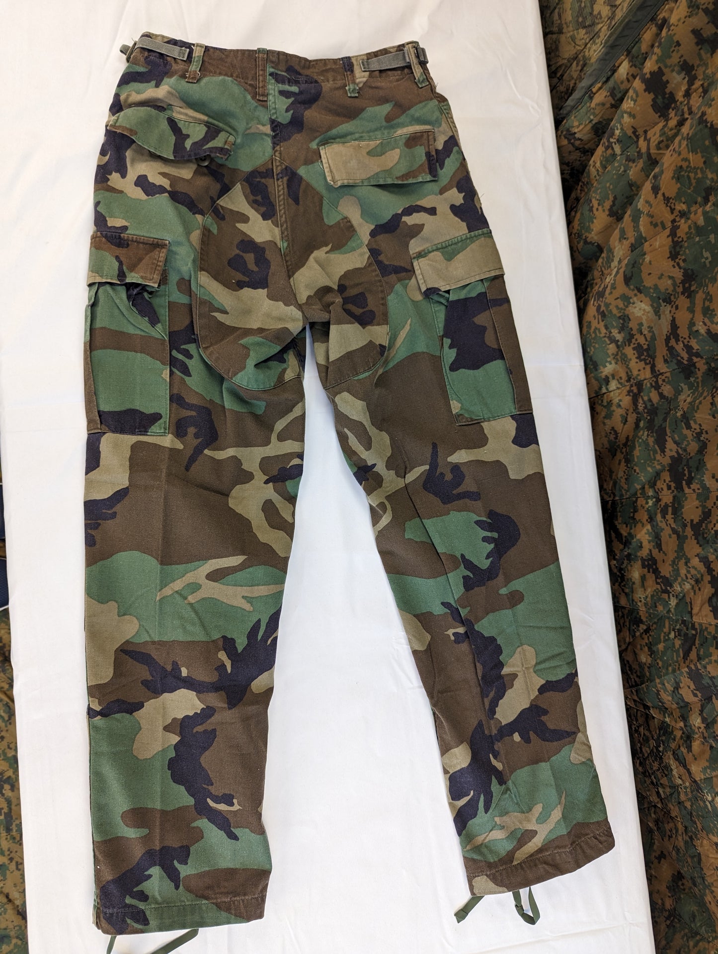 A full view of a pair of U.S. Military Surplus Combat Trousers in Woodland camouflage, laid flat on a white background, displaying the cargo pockets, knee reinforcement, and drawstring cuffs.