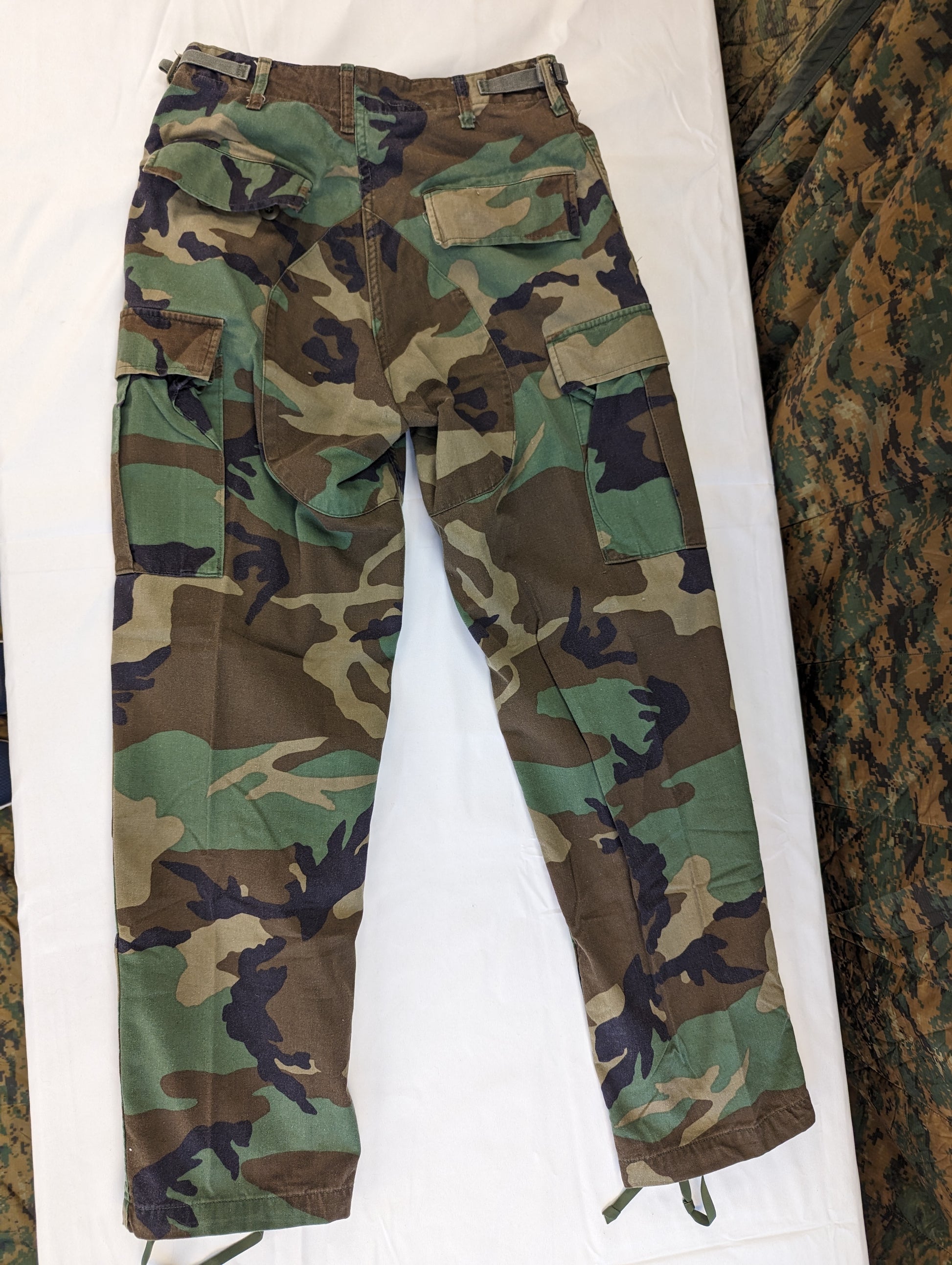 A full view of a pair of U.S. Military Surplus Combat Trousers in Woodland camouflage, laid flat on a white background, displaying the cargo pockets, knee reinforcement, and drawstring cuffs.