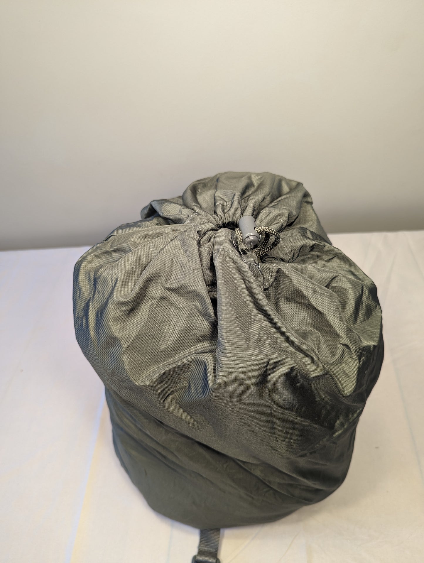A large, tightly compressed U.S. Military Surplus Compression Stuff Sack in olive green, standing upright, featuring a cinched drawstring closure and a wrinkled nylon texture.